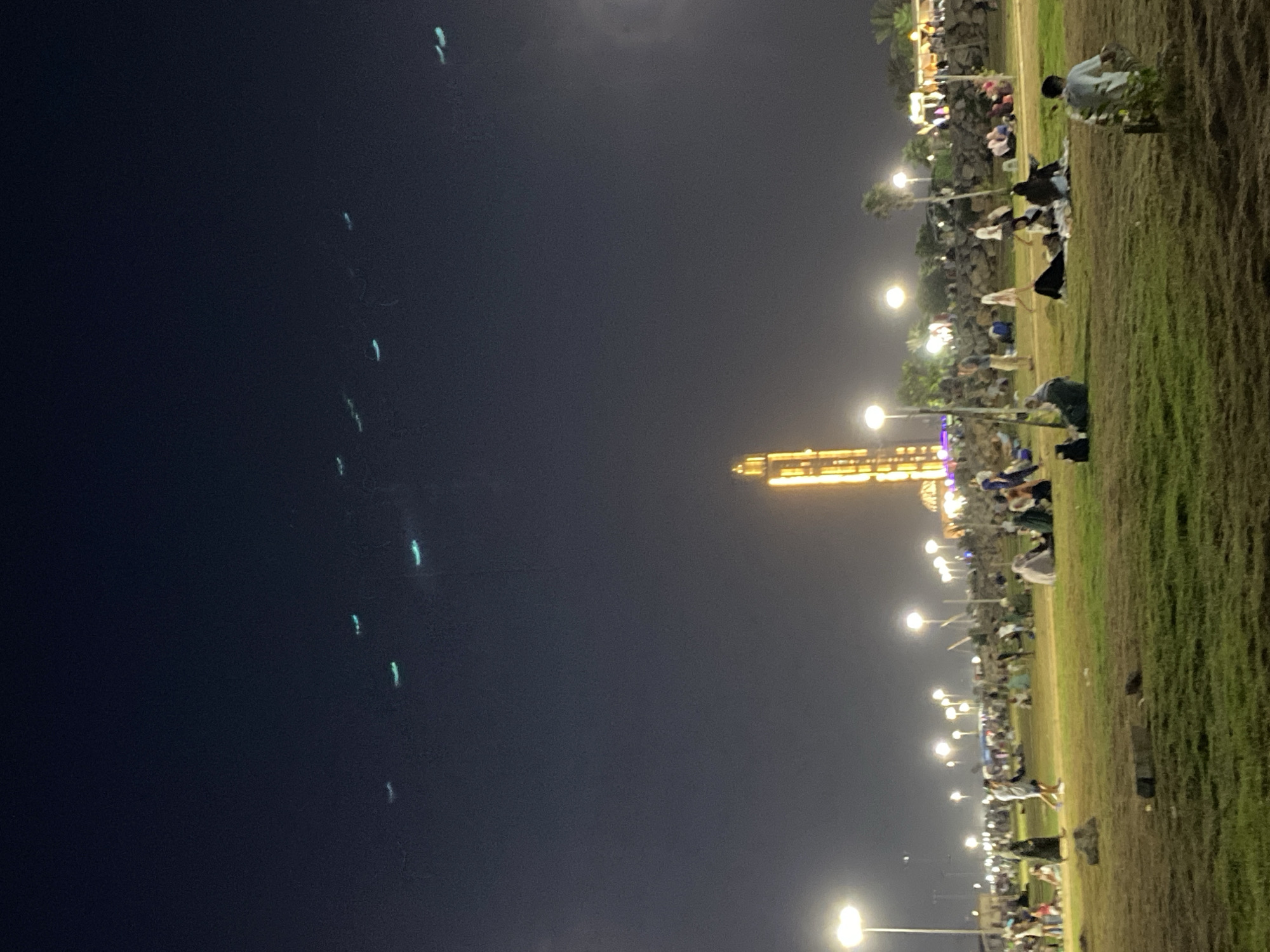 The image depicts an evening scene at a park in Hussein Dey, Algeria, under a dark sky illuminated by artificial lights. In the foreground, a grassy area is dotted with rocks and a few small plants. Several people are scattered across the grass, some sitting in groups and others walking. The middle ground features a pathway lined with lampposts casting bright pools of light. Beyond the pathway, a crowd of people is visible, congregating near structures that appear to be part of an event or attraction. A prominent, brightly lit tower dominates the background, its golden light piercing the darkness. High in the sky, a series of faint, turquoise lights form a linear pattern, suggesting aircraft or perhaps a drone show. The overall atmosphere is one of a public gathering, with an emphasis on light and activity in the urban park setting.