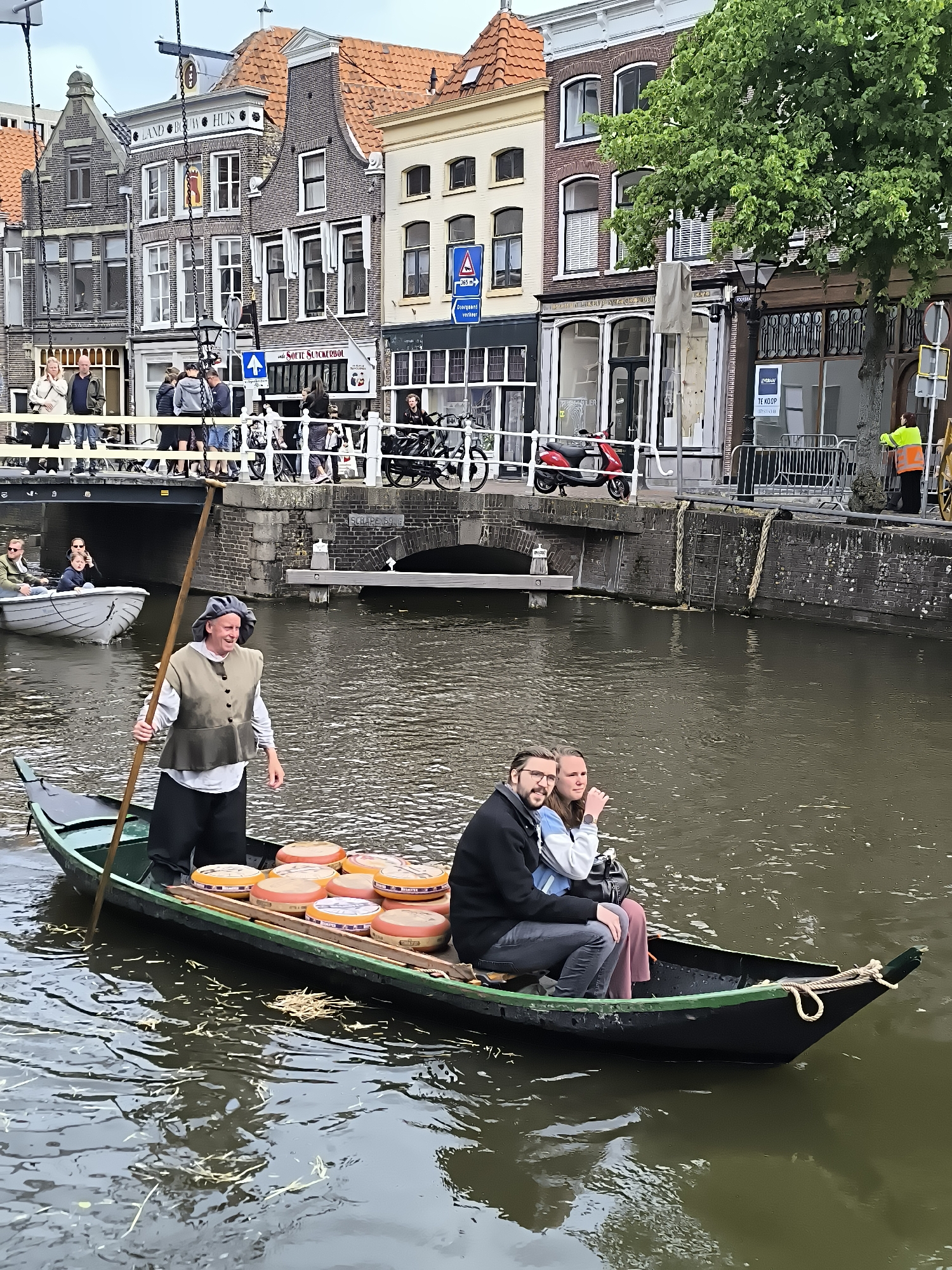 This image captures a vibrant canal scene in Alkmaar, Netherlands.
In the foreground, a dark green, narrow boat, approximately 5-6 meters in length, navigates the canal. A male individual stands in the bow, dressed in a white long-sleeved shirt, a dark olive-green sleeveless vest with brass buttons, dark trousers, and a grey, soft, rounded cap. He holds a long wooden pole vertically, partially submerged in the water, propelling the boat. He smiles, looking towards the left side of the frame. Behind him, approximately ten large, round, orange-colored cheese wheels are arranged on wooden planks within the boat, suggesting a cheese market or traditional delivery. In the stern of the same boat, a male passenger with a beard and glasses, wearing a dark jacket, sits beside a female passenger wearing a light blue and white patterned top and dark trousers. Both are looking towards the front/right, exhibiting slight smiles.
The canal water is dark, showing ripples and reflections, with scattered light-colored organic debris floating on its surface. Further back and to the left, a smaller white rowboat contains two individuals: an adult male at the oars and a child in the front.
A stone arch bridge crosses the canal in the mid-ground, with the inscription "SCHAPE G." partially visible on its side. Numerous casually dressed pedestrians are gathered on this bridge, observing the canal below.
The background features historic brick buildings lining both sides of the canal, characterized by distinct Dutch gable roofs (stepped and bell gables) and numerous windows, many with white frames. Prominent storefronts include "LAND BOUW HUIS" (Agricultural House) on a light-colored facade and "SOETE SUCKERBOL" (Sweet Sugar Loaf) on a darker building. Street-level details on the canal bank include white railings, a parked red scooter, various street signs (one indicating a "2.65 m" height restriction and another a blue directional arrow), and metal barricades. A person wearing a high-visibility yellow jacket stands near a tree and barricades on the far right bank. Lush green trees are present along the canal, with one partially obscuring a building facade on the right. The sky is overcast and bright, indicating diffused daylight.