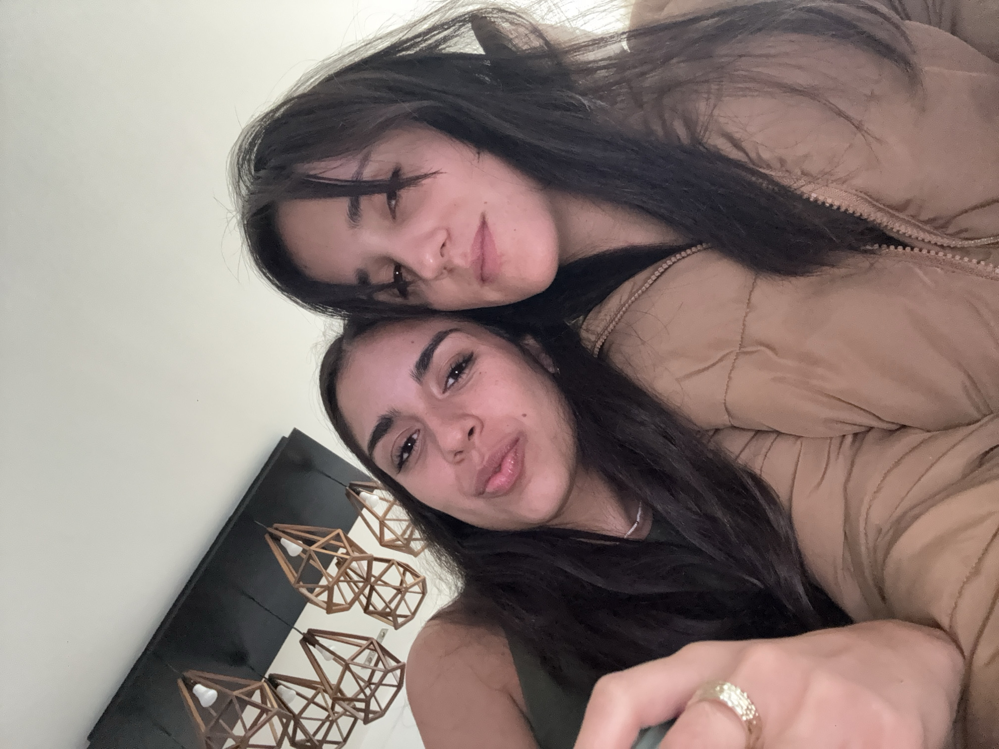 Two young women are posing for a selfie. One is in the foreground, wearing a green tank top and a ring on her right hand. The other woman is behind her, wearing a brown puffer jacket. Both women have dark hair and are looking at the camera. Behind them is a white wall with a black rectangular panel mounted on it. The panel has several geometric wooden lamps hanging from it. Based on the style of the lamps and general aesthetic, the location is likely in a modern home or apartment. However, the image does not provide enough information to definitively determine the location within Mexico City.