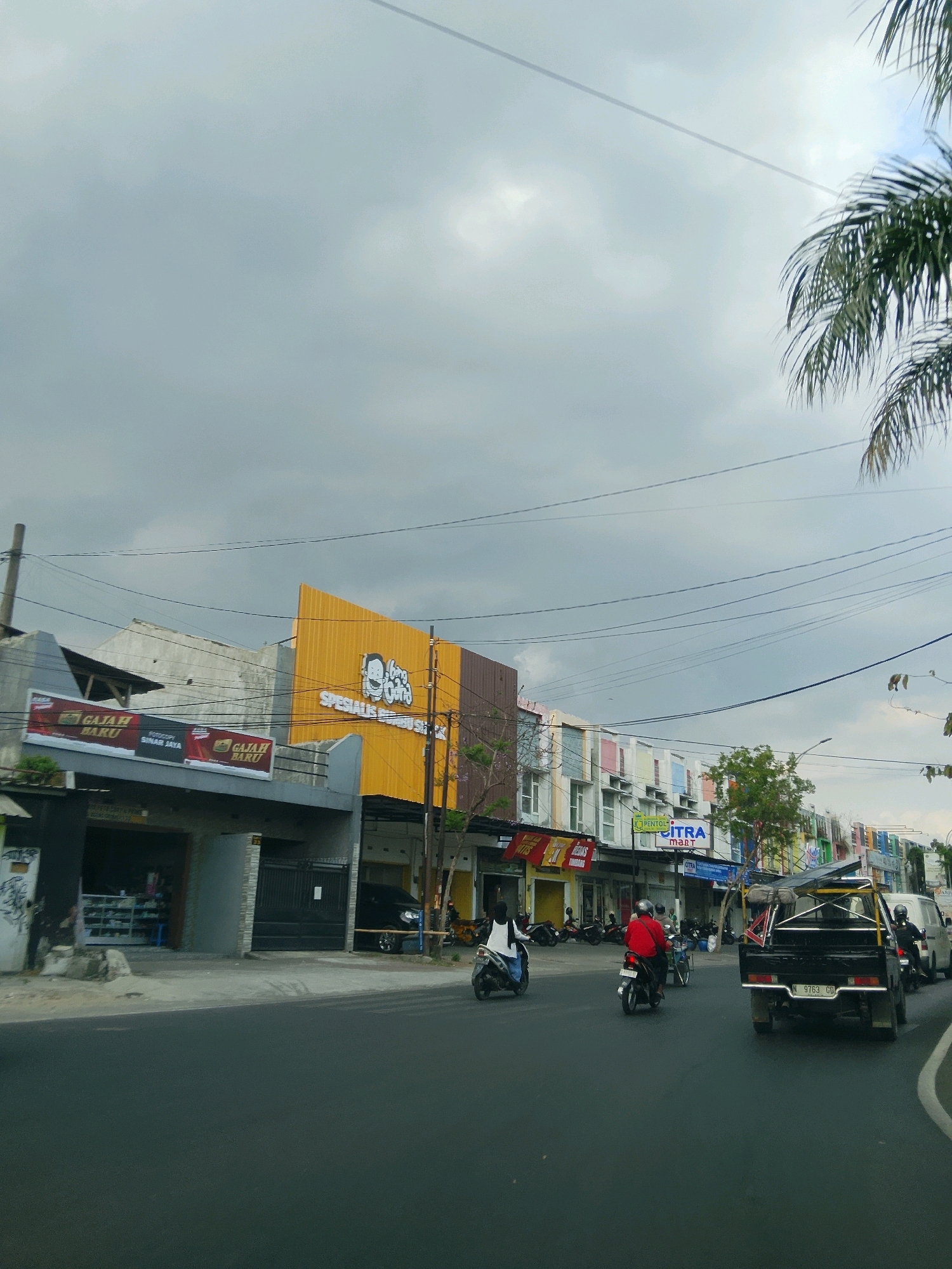 The image depicts a street scene in Malang, Indonesia, under an overcast sky. A two-lane asphalt road runs horizontally across the foreground, with a slight curve to the right. Several vehicles are present, including two motorcycles in the left lane, one carrying a rider in white, and another with a rider in red. In the right lane, a black pickup truck with a roof rack is driving away from the camera, followed by a white van. The buildings lining the street are mostly two to three stories high, with a variety of facades and signage. On the left, a yellow building features a cartoon character and text that reads "SPESIALIS BAKSO". Adjacent to it are other businesses with visible signage, including "GAJAH BARU". Further down the street, a row of buildings displays colorful window frames. Overhead, a network of electrical wires crisscrosses the sky, and palm fronds from a tree are visible in the upper right corner. The overall impression is that of a bustling commercial street in an Indonesian city.