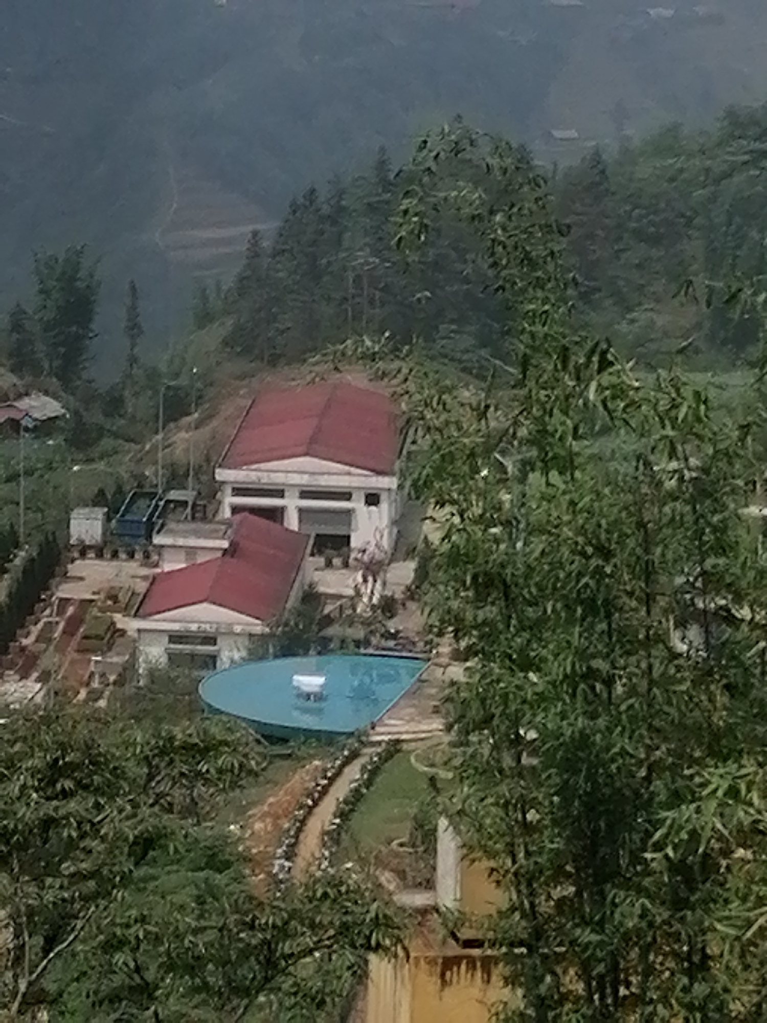 The image depicts a scene in Sa Pa Ward, Vietnam. A building complex with red roofs is visible, situated amidst lush greenery. A circular pool with a fountain feature is located adjacent to one of the buildings. Trucks or other vehicles are parked near the building complex. Trees and vegetation dominate the foreground and background, partially obscuring the view. In the distant background, terraced land can be discerned. The overall scene suggests a mountainous or hilly terrain.