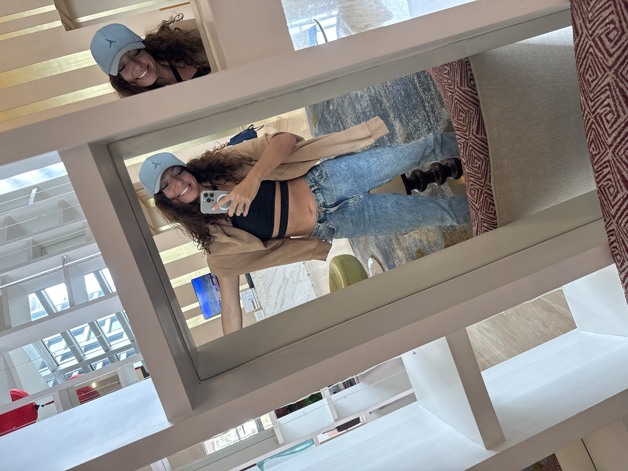 The image shows a woman reflected in a large mirror. She is wearing a light blue baseball cap, a black crop top with two straps, light-wash blue jeans, and a tan jacket. She's holding a phone and smiling at her reflection. Another reflection of the woman is visible at the top of the mirror, also smiling. The mirror is part of a large, white, shelving unit structure. Behind the main reflection, there is a partial view of an interior space including a TV, a green chair, and a rug. Outside of the windows visible in the top left of the image, there appears to be an overhead view of Đà Nẵng, Vietnam based on the distinctive architectural elements such as the city's bridges and the overall urban layout that can be observed.