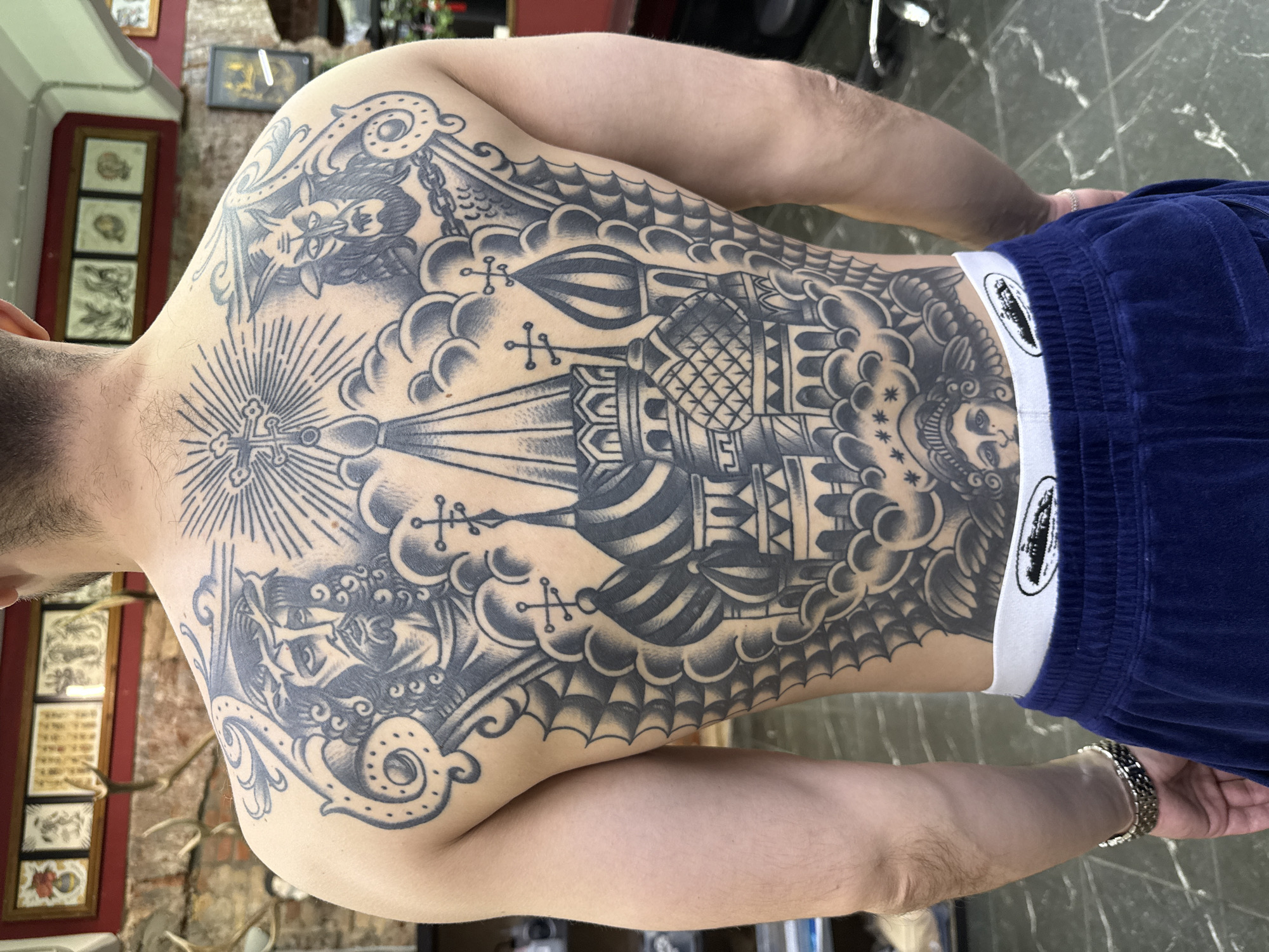 A person is seen from the back, revealing a large, detailed black and gray tattoo covering their entire back. The tattoo features a depiction of Saint Basil's Cathedral, an Orthodox church located in Red Square, Moscow, Russia. Additional elements in the tattoo include religious imagery like crosses, winged demonic figures, and ornate patterns resembling clouds and decorative motifs. The person wears blue pants and a white waistband is visible. The background includes a glimpse of a room with decorative items and framed artwork on the walls, and a patterned floor.