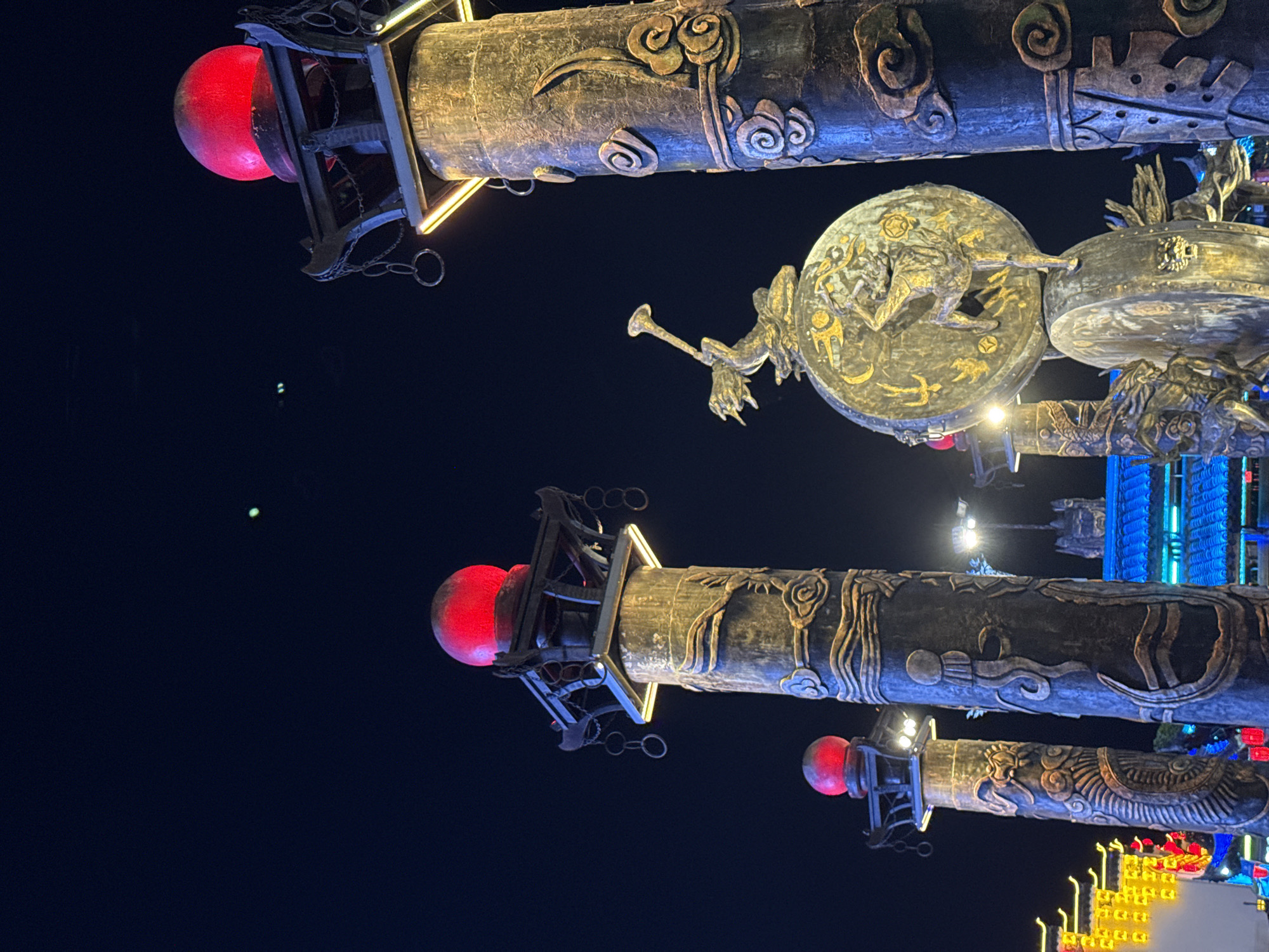 The image presents a striking, upward-angled view of highly ornate and illuminated architectural structures at night in Wulingyuan, China. The scene is dominated by several large, dark, cylindrical beams or pillars that are richly decorated with intricate, metallic-looking carvings in gold and bronze tones. These carvings feature stylized cloud patterns, swirling motifs, and complex designs that suggest mythical creatures or ancient symbols.
Prominently featured are several bright, glowing red spherical lights, housed within dark, cage-like frames, attached to the ends of some of the larger structures. One particularly detailed element is a large, circular disc-shaped ornament positioned centrally, depicting a dynamic figure, possibly a deity or mythical hero, surrounded by smaller symbolic patterns. A smaller, similarly decorated cylindrical structure with a red light is also visible towards the bottom right.
The background is a deep, dark night sky, indicating the image was captured after sunset. In the distance, various artificial lights are discernible, including prominent blue vertical lights and clusters of yellow/orange lights, suggesting other illuminated buildings or features within the complex. There is no human activity taking place; the focus is solely on these elaborate, decorative structures. No legible text is visible within the image. The overall impression is one of traditional grandeur and intricate craftsmanship under the night sky.
