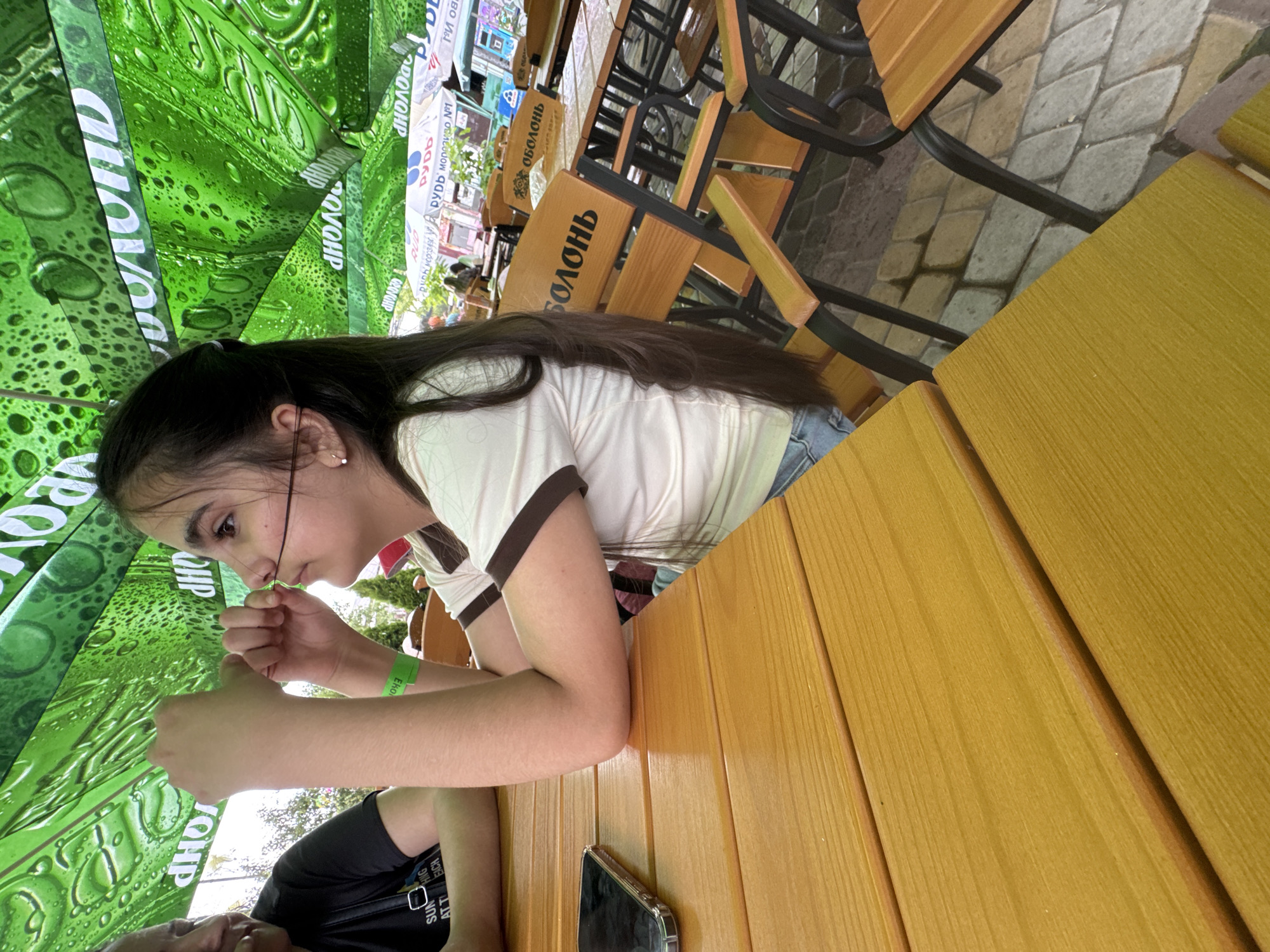 In Zaliznychne, Ukraine, under a green, droplet-patterned umbrella at an outdoor seating area, a young girl with long, dark hair is focused on manipulating a thin black string between her fingers and nose. She is wearing a white shirt with brown trim and a green wristband. To her left, another person is partially visible, wearing a black t-shirt. The scene is set at a wooden table, with a smartphone resting on it. In the background, several wooden chairs with black metal frames are arranged around tables, and the ground is paved with cobblestones.