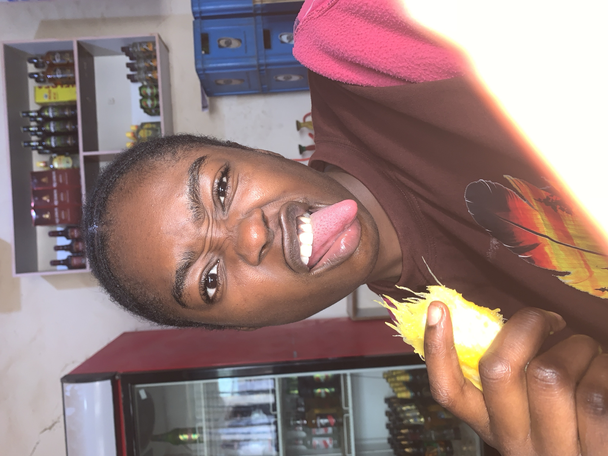 A person is holding a piece of yellow fruit while sticking their tongue out in a store. The person has dark skin, short black hair, and is wearing a brown shirt with a pink collar. The fruit is irregularly shaped with stringy yellow protrusions. In the background, there are shelves with bottles of liquor and blue crates, and a red and white refrigerator stocked with drinks. The location is the city of New Stadium, Nigeria.