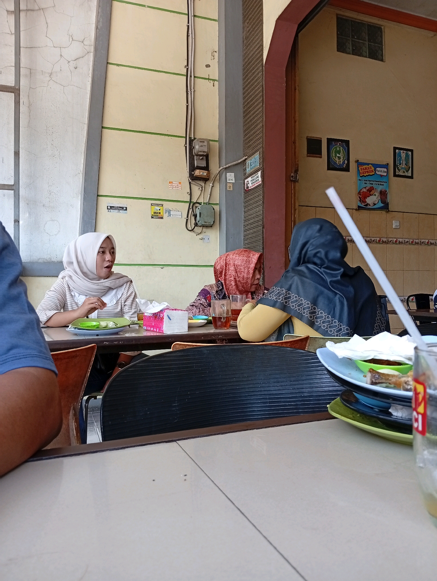 The image depicts three women seated at a table, likely in a restaurant in Kongsijaya, Indonesia. Two of the women are wearing hijabs. The woman on the left, wearing a light-colored striped shirt and a white hijab, is looking to her right and appears to be mid-conversation. She has a plate with food and a tissue box in front of her. The woman in the middle, wearing a red and patterned hijab, is looking down at her plate. The woman on the right is wearing a dark hijab and a yellow shirt. She is facing away from the camera and appears to be looking at something to her right. The table is set with plates, glasses of tea, and possibly some food remnants. In the background, there are posters on the wall, an electrical meter, and some architectural features of the building. The scene suggests a casual meal or gathering.