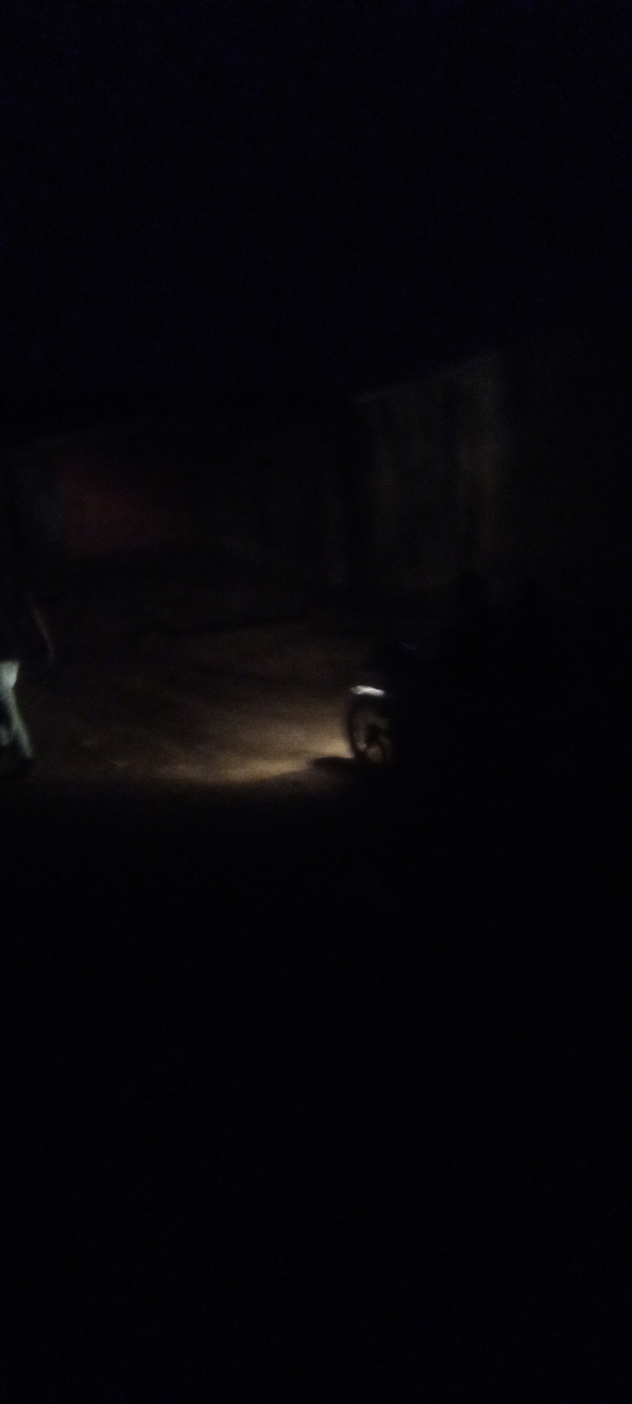 A dimly lit scene in Sabon Tasha, Nigeria, features a figure walking toward a stationary motorcycle. The motorcycle has its headlight on, illuminating the ground in front of it. A building is visible in the background. The overall environment is dark and sparsely illuminated.