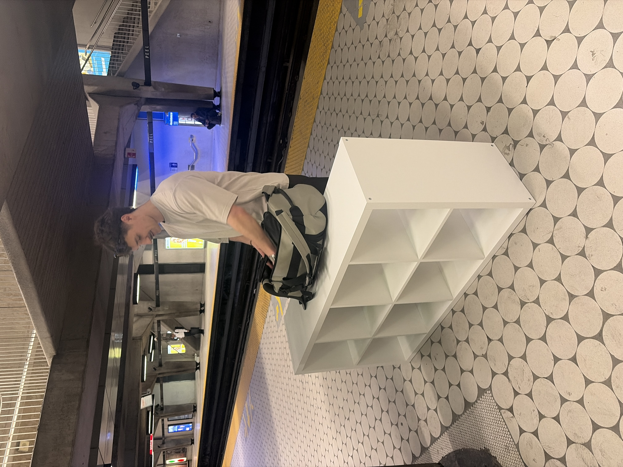 The image captures a scene within an underground subway station, featuring a young man and a large white storage unit in the foreground. The man, with dark, curly hair, wears a light-colored t-shirt and light-colored pants, and carries a gray and black backpack on his back. He is looking downwards, possibly adjusting his backpack or interacting with the white shelf unit. This prominent object is a cubby organizer with multiple square compartments.
The station platform is characterized by a distinctive floor pattern of large, light-colored interlocking ovals on a darker base, transitioning to a smaller mosaic tile pattern in one area. A yellow textured warning strip runs along the edge of the platform next to the subway tracks. Concrete pillars and artificial overhead lighting are visible, typical of a metro environment. In the background, other indistinct figures can be seen on the platform.
Visible text on a black vertical pole clearly reads "PEEL," indicating the location is the Peel metro station in Montreal, Canada. Another sign in the distance also appears to bear the "PEEL" name. The activity is generally passive, showing passengers on a platform, with the large white shelf unit being a notable, somewhat incongruous detail within the setting. The time of day is not determinable due to the indoor, artificially lit environment.