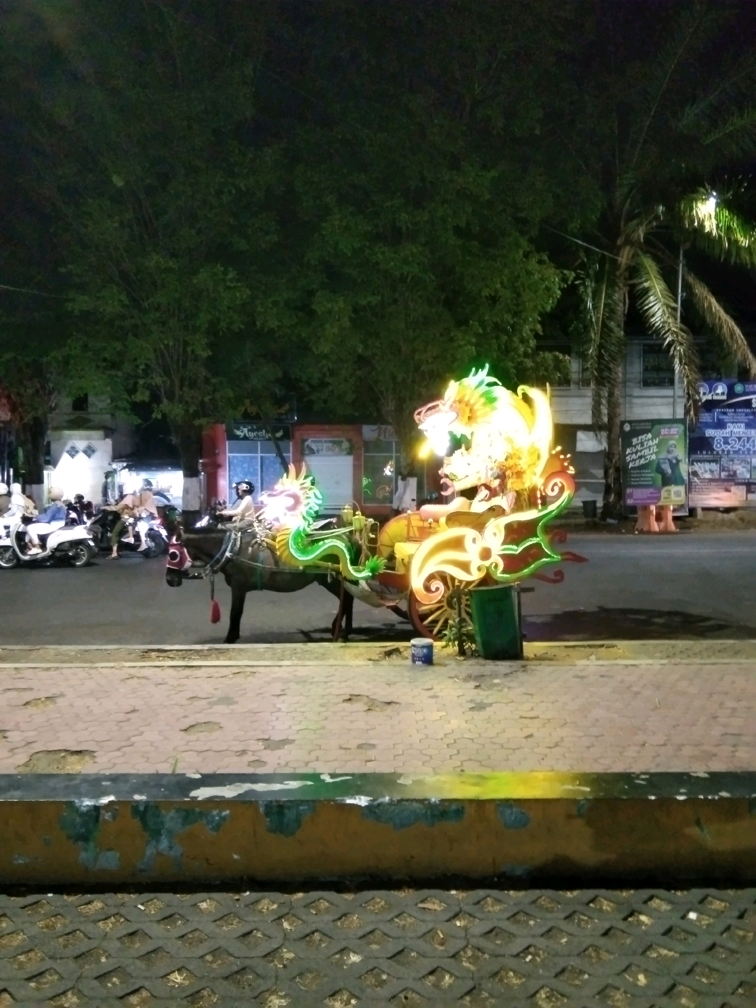 In Bondowoso, Indonesia, at night, a horse-drawn carriage is illuminated by vibrant, neon dragon-shaped lights. The carriage is decorated with colorful elements and appears to be parked or slowly moving along a street. A person is seated in the carriage, likely the driver, wearing a helmet. In the background, several motorbikes are visible, some with riders. The environment suggests a public area with trees and buildings lining the street. The ground is paved with interlocking bricks, and there is a low concrete barrier in the foreground. The overall scene depicts a festive or recreational atmosphere at night in a city setting.