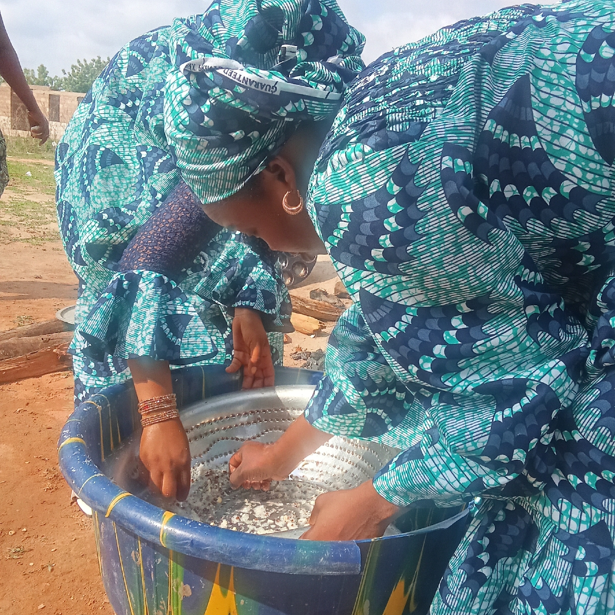 Two women in matching blue and green patterned dresses and headwraps are bent over a large, blue tub with yellow stripes, washing a grain-like substance. One woman wears gold hoop earrings and bracelets on her wrist. The location appears to be outdoors, in a dirt area. A building is visible in the background. The word "GUARANTEED" is visible on the headwrap of one of the women.