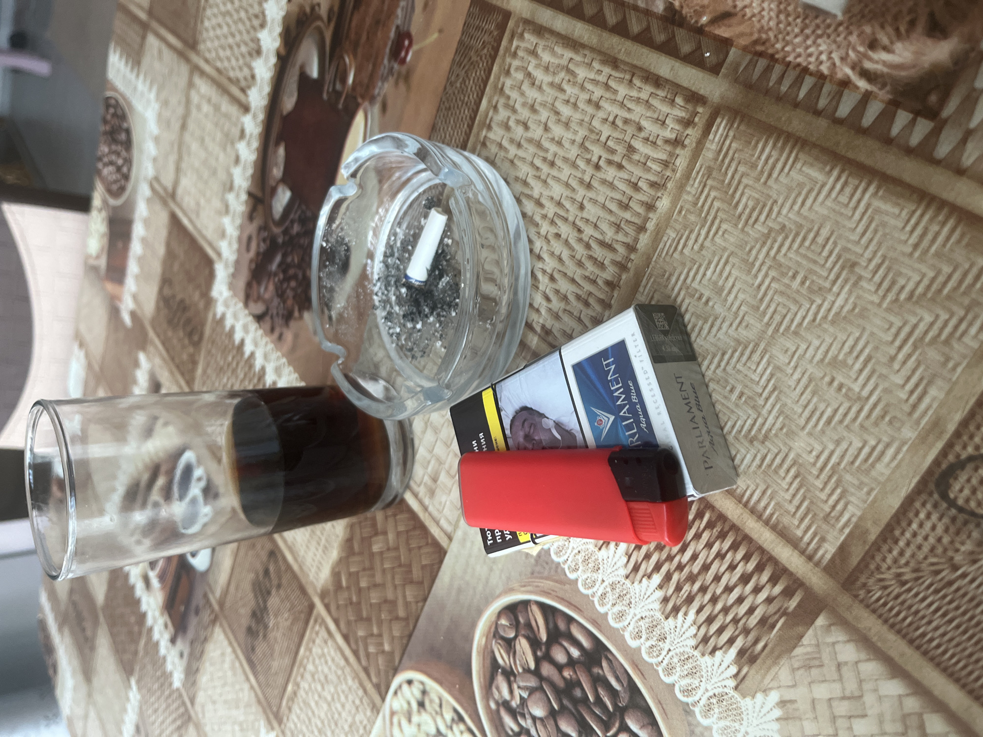The image captures a tabletop scene, likely in Novi Pazar, Bulgaria, featuring a collection of items associated with a coffee and smoke break. A clear glass, partially filled with a dark liquid resembling coffee, stands on the left. To its right, a transparent glass ashtray contains a white cigarette butt and a considerable amount of ash, indicating recent use. Adjacent to the ashtray lies a pack of "Parliament Aqua Blue" cigarettes, with a bright red lighter placed on top of it.
The table is covered with a patterned tablecloth, which features sections adorned with coffee beans and the word "Coffee" inscribed within them, suggesting a coffee-themed setting. The cigarette pack prominently displays a health warning, showing an image of a person connected to medical equipment. Visible text on the pack includes "PARLIAMENT Aqua Blue," "ALL RECESSED-F," and partially legible Cyrillic text, such as "ПРЕДУПРЕЖДЕНИЕ:" (Warning:). The lighting suggests it is daytime. No individuals are visible in the frame, but the arrangement implies a personal moment of relaxation.