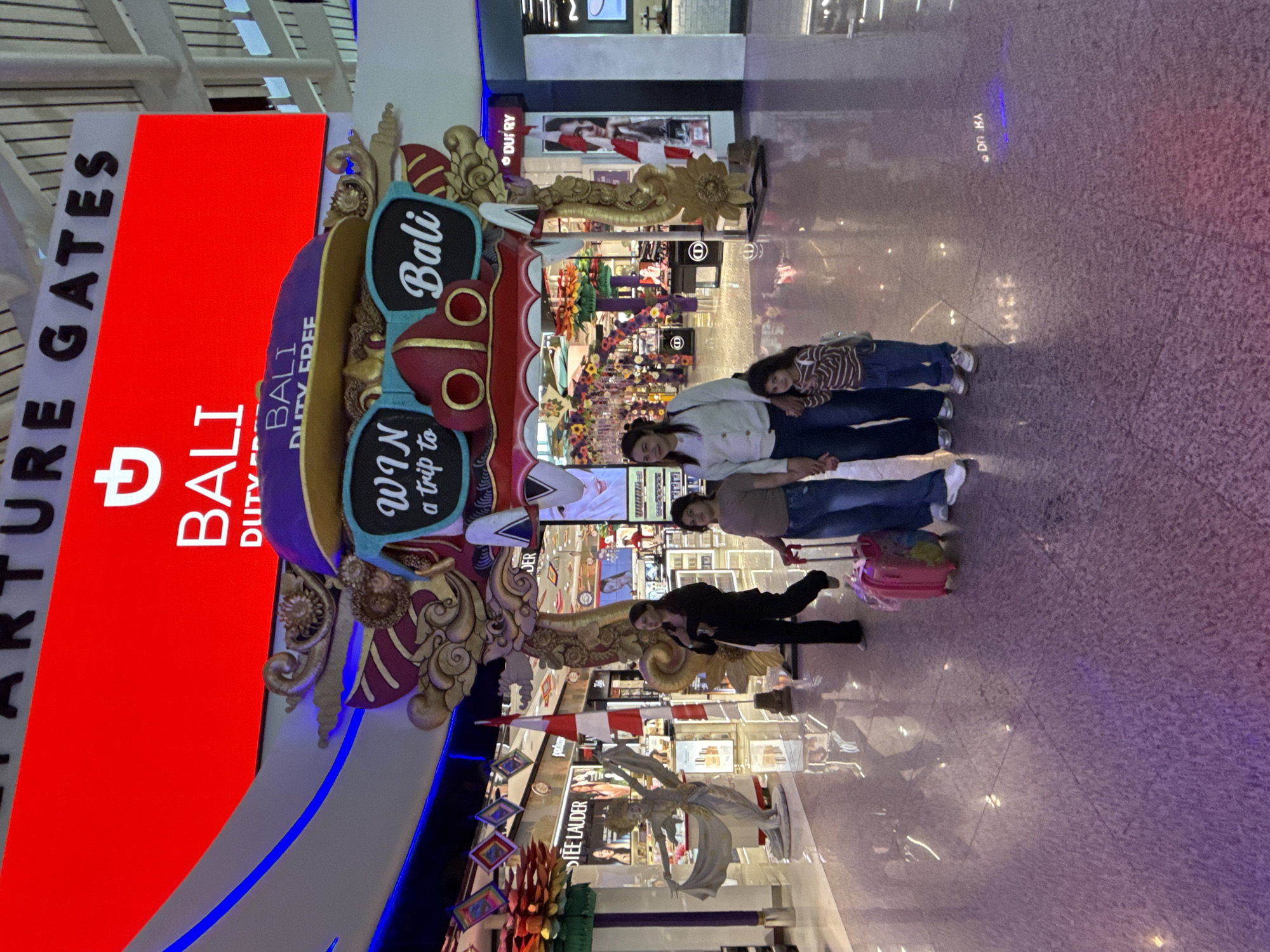 In Kuta, Indonesia, inside an airport's departure area, a family of four is walking through a brightly lit walkway. A woman, holding the hand of a young girl, is dressed in a white sweater and blue jeans. The young girl wears a striped shirt and blue jeans. Behind them, another woman is walking with a pink suitcase. The environment is adorned with a large, colorful archway featuring an artistic representation of sunglasses with the words "WIN a trip to" and "BALI DUTY FREE." Above this, a large red LED screen displays "DEPARTURE GATES" and "BALI DUTY FREE." Shops line the background, with displays of beauty products and signage, including a prominent "ESTEE LAUDER" store. The floor is a polished, reflective surface.
