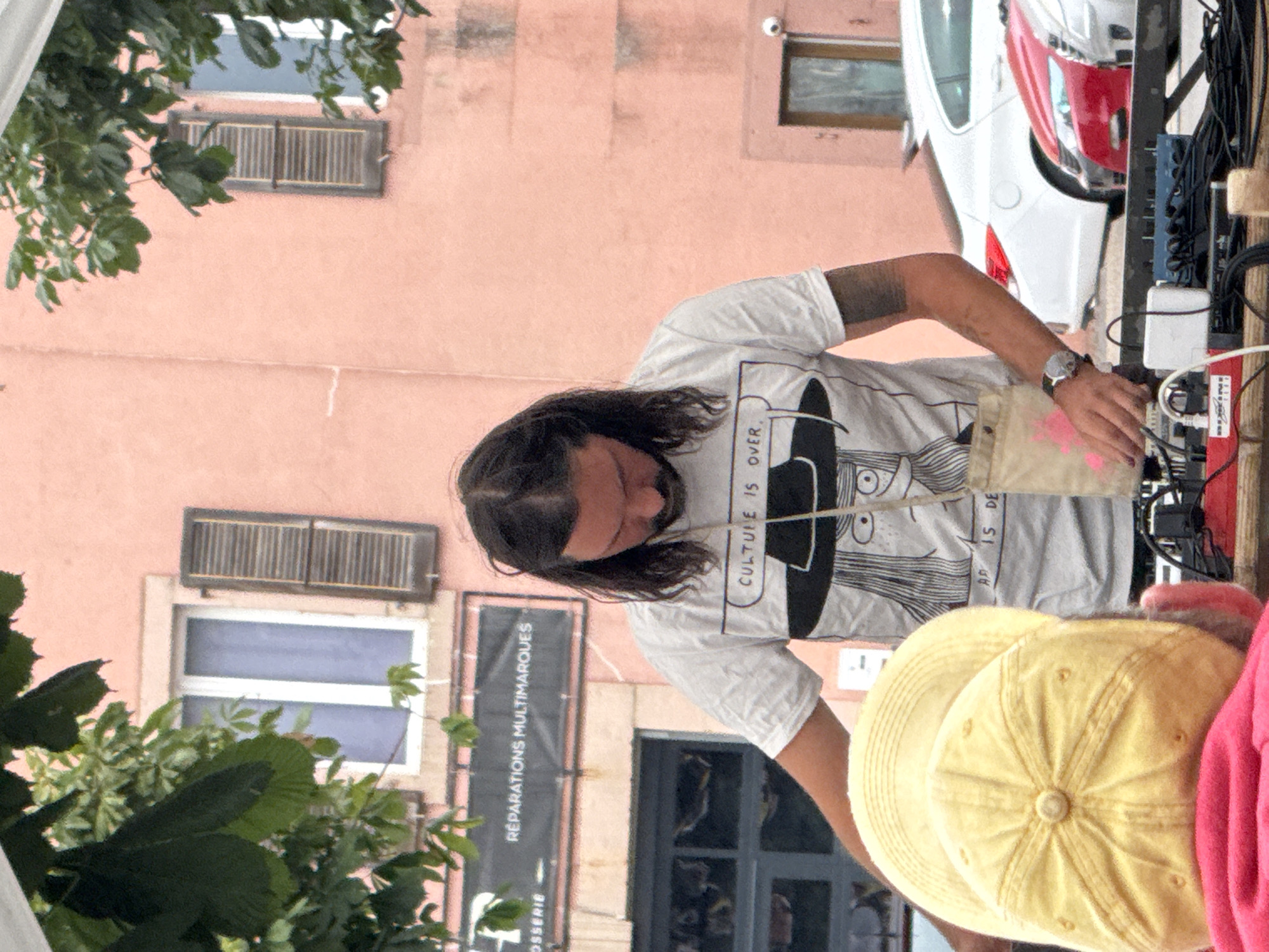 In La Chaux-de-Fonds, Switzerland, a man with long dark hair and a beard stands outdoors, looking down at a table of electronic equipment. He is wearing a white t-shirt with a graphic print of a person wearing a hat, and the text "CULTURE IS OVER". A canvas bag with a pink splatter design hangs from his neck. In the foreground, a person wearing a yellow baseball cap and a pink shirt is visible from behind. The background shows a salmon-pink building with windows and dark shutters, and parked cars, including a white sedan and a red car. There is also signage on the building that reads "RÉPARATIONS MULTIMARQUES". Green foliage hangs from trees above.