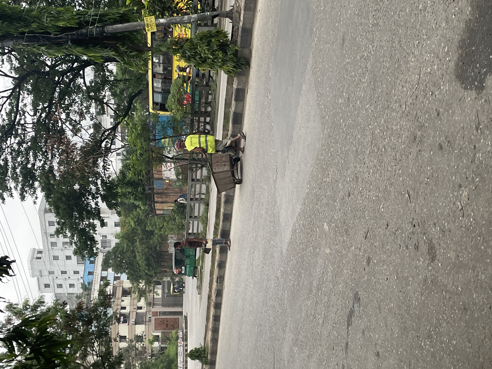 The image, taken in the city of Dhaka, Bangladesh, depicts a street scene. A man in a yellow safety vest is pulling a metal cart along the side of the road. Another man is visible walking on the sidewalk near a green vehicle. Buildings, trees, and other vehicles are present in the background. The street is made of asphalt. The overall environment suggests an urban setting.