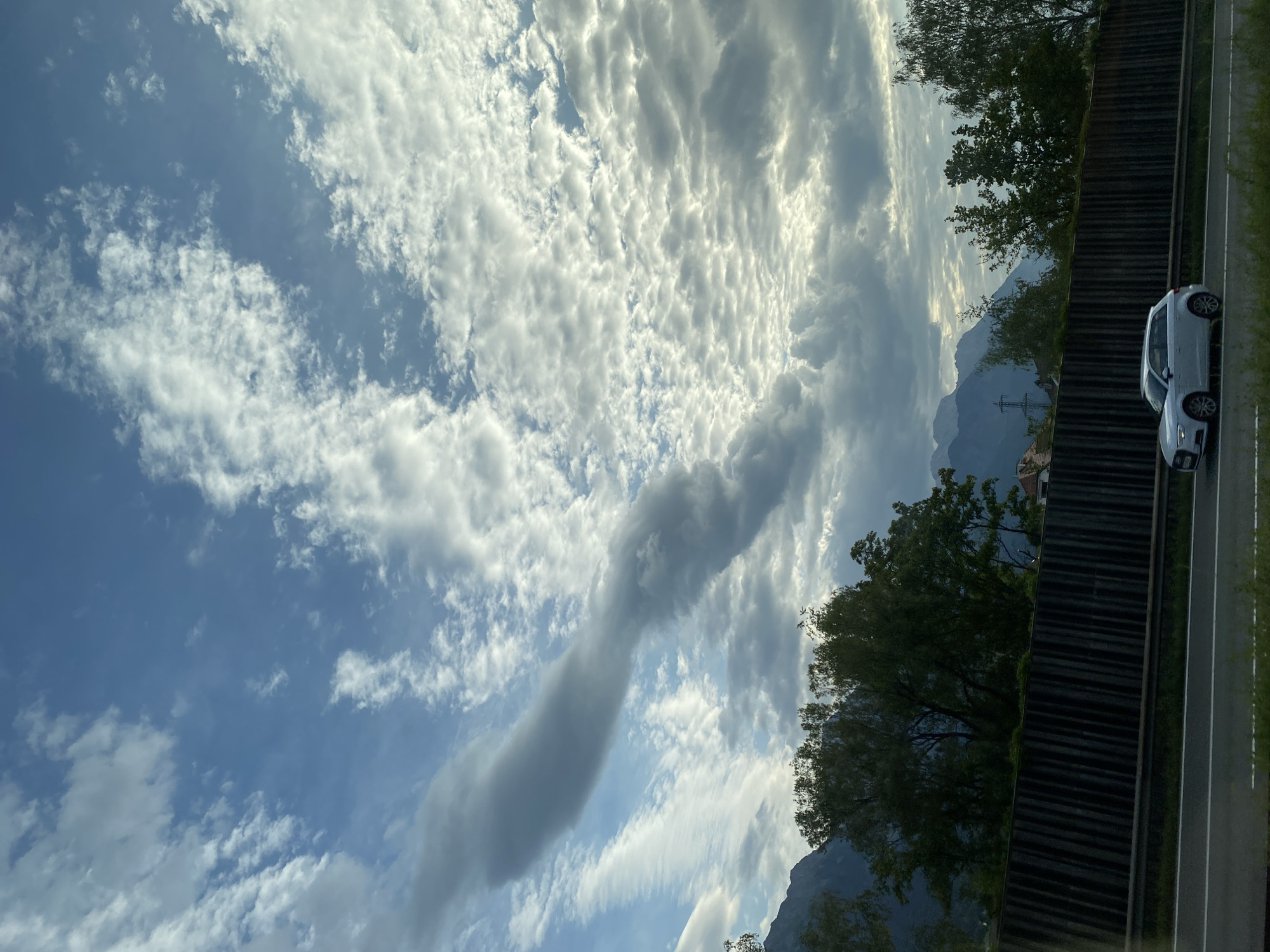 This media file captures a scenic view from a roadside, likely on a highway near Innsbruck, Austria. The primary subjects are the sky, mountains, and a passing white car. The sky is dramatic and filled with a variety of clouds, including wispy cirrus formations and larger, textured stratocumulus clouds that create a rippled appearance. A particularly striking, elongated cloud formation is visible in the center-left of the frame. The weather appears to be partly cloudy, with sunlight breaking through the clouds in places, suggesting it's either morning or late afternoon.
In the background, rugged mountains are partially obscured by trees and the clouds, hinting at a mountainous landscape. The foreground features a paved road with white lane markings, a dark, textured barrier, and lush green trees lining the edge. A modern white car is captured in motion, traveling on the road alongside the barrier. The overall scene evokes a sense of travel and natural beauty. There is no visible text in the image.