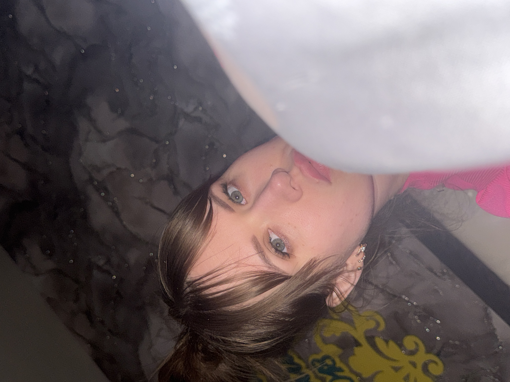 A close-up portrait features a young woman with a fair complexion, striking green-blue eyes, and light brown hair, which falls across her forehead and shoulders. She is wearing pink lipstick and a pink garment, possibly a top, is visible on the right side of the frame. Her left ear is adorned with multiple small, sparkling earrings. The image is taken from a tilted perspective, giving the impression that she is lying down or viewed from a low angle.
The background primarily consists of a dark, reflective surface, possibly a floor or table, which exhibits a subtle, wavy pattern and numerous small, circular light reflections, suggesting glitter or bokeh. In the lower right corner, a section of a yellow and green patterned fabric or surface, resembling a rug or upholstered item, is visible. The scene appears to be indoors. The woman is looking directly towards the camera with a neutral and calm expression. No specific activity is depicted other than posing for the photograph. There is no visible text in the image. The location context for this image is Chaikovsky, Russia.