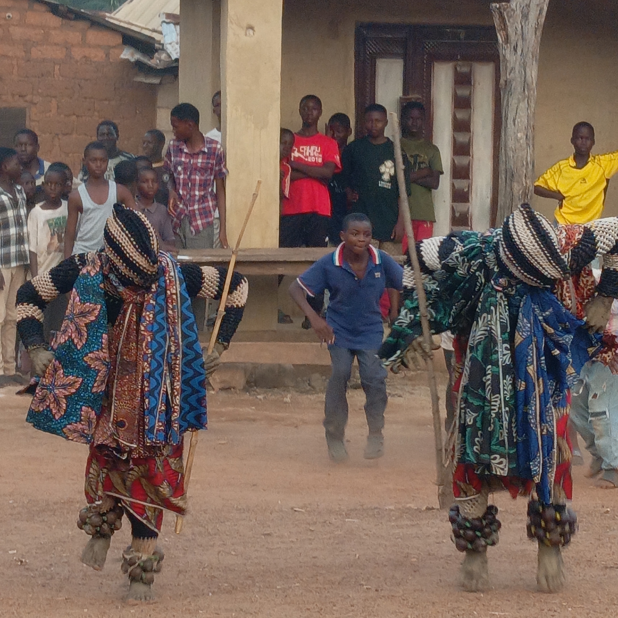 The image depicts an outdoor scene in Akwanga, Nigeria. Two figures dressed in elaborate, colorful costumes dominate the foreground. They appear to be performing or participating in a traditional ceremony, each holding a stick. On their legs they have clusters of what appear to be fruit or other natural items. Behind them, a group of young men and boys are gathered, observing the event. One boy in a blue shirt is jumping. The location appears to be a village setting with simple buildings in the background. The ground is dirt or packed earth. The general impression is that of a cultural performance or celebration.