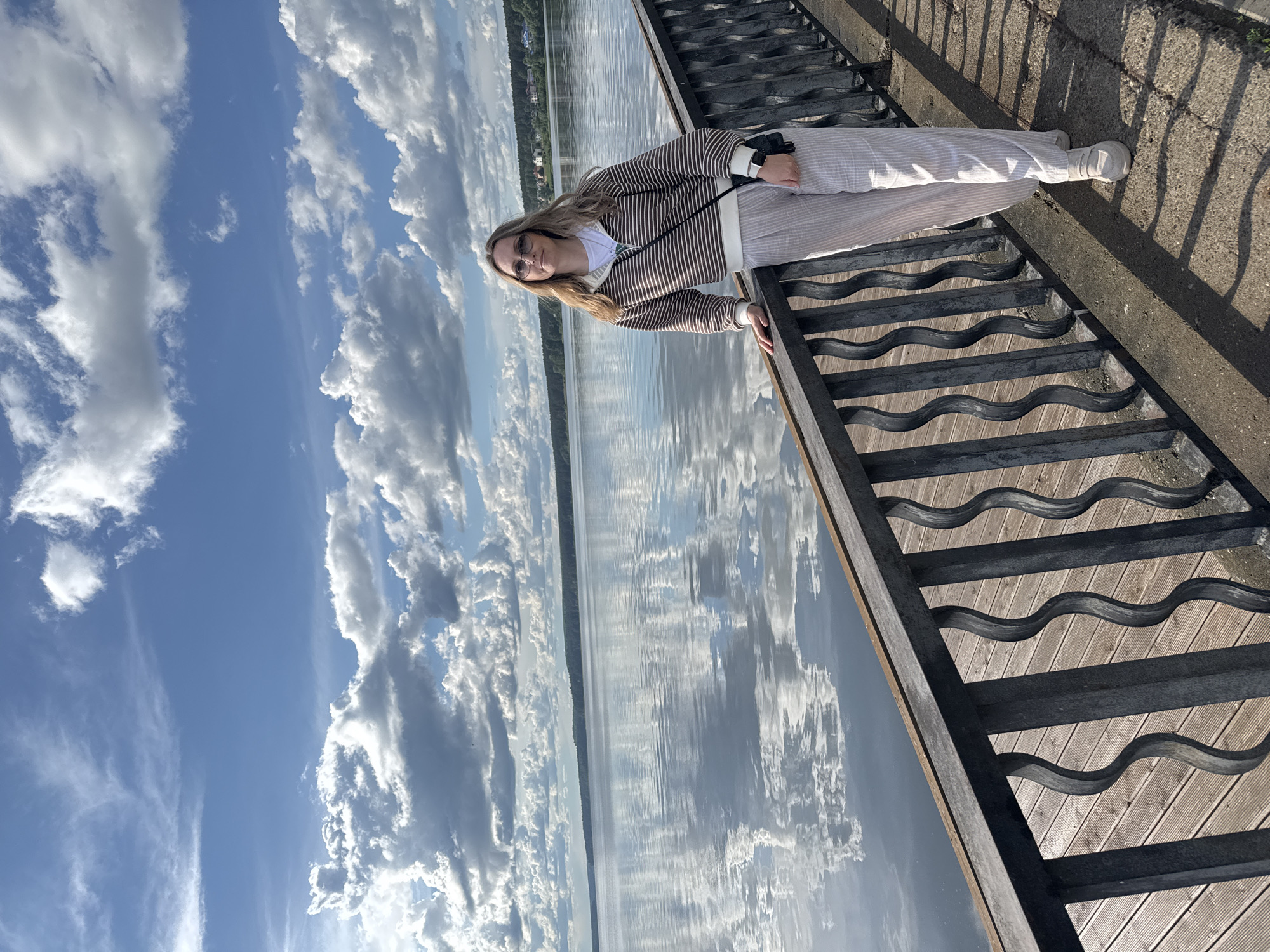A woman is standing on a wooden walkway next to a body of water. She has blonde hair and is wearing glasses, a striped brown and white jacket, and light-colored pants and shoes. She is holding onto the railing of the walkway. The water reflects the sky, which is blue with white clouds. The walkway appears to be part of a dock or pier. In the distance across the water, there is a treeline. This is likely the Votkinsk Reservoir on the Kama River in the city of Votkinsk, Russia.