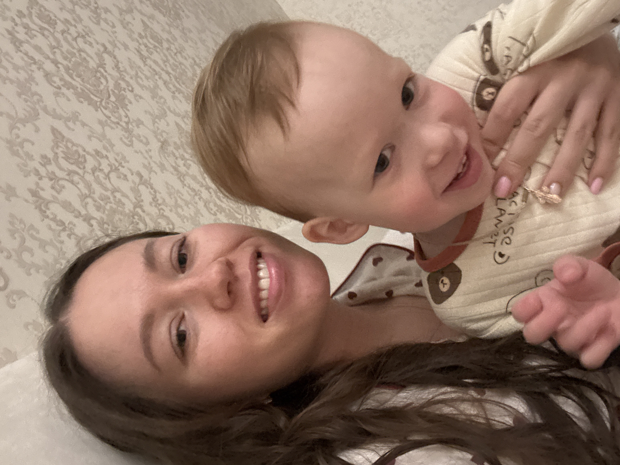 The image shows a selfie of a woman with a young child. The woman has brown hair and is smiling. The child has blond hair and is also smiling, wearing a white pajama top with a brown bear print. The background appears to be a textured wall, possibly wallpaper. The scene likely takes place indoors, possibly a bedroom, though the location of Ferma, Russia is not discernible in the image.