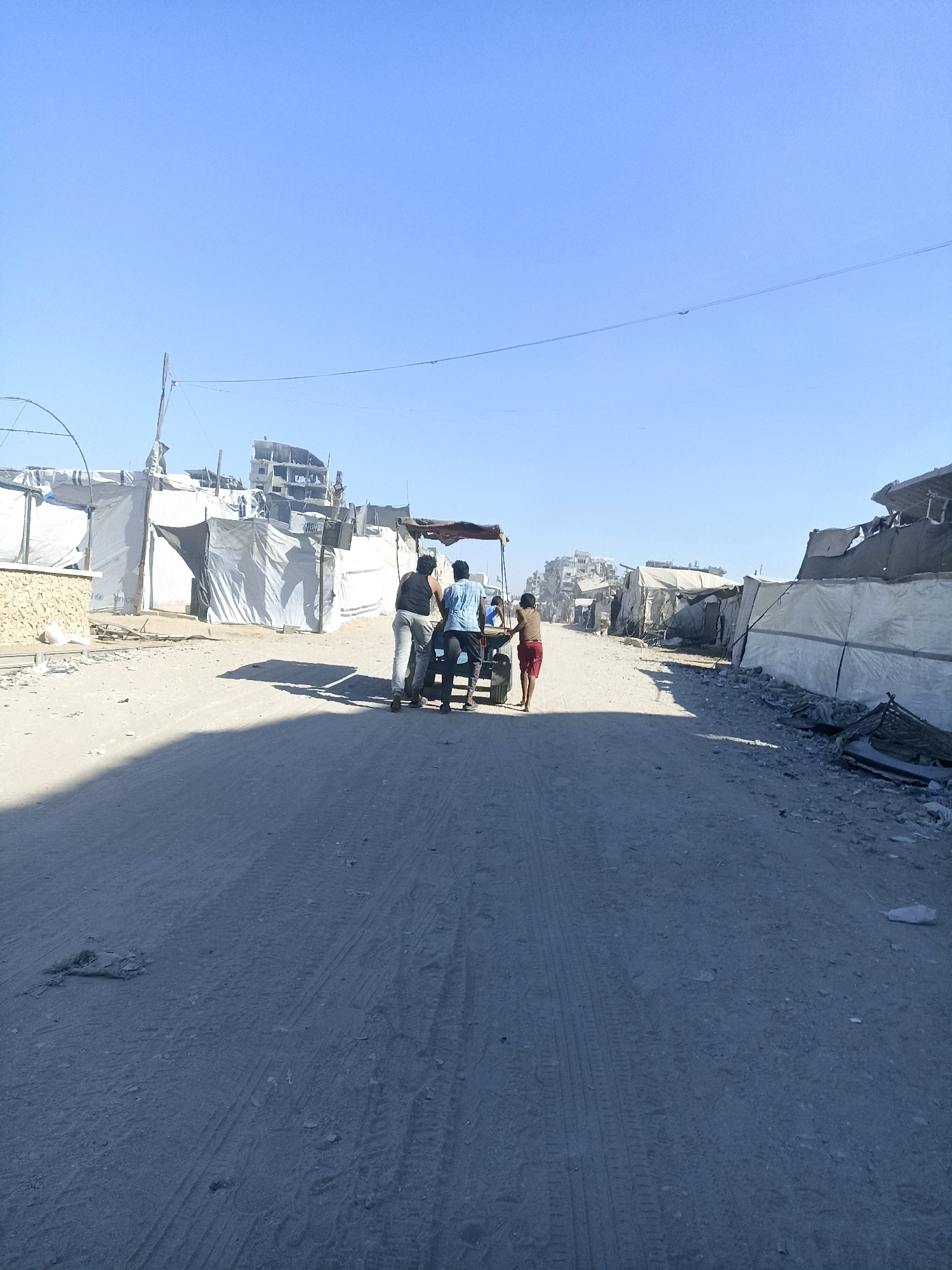 The image depicts a street scene in what appears to be a temporary settlement or a damaged urban area in Gaza, Palestinian Territories. The ground is unpaved, dusty, and shows tire tracks, suggesting it is a road or pathway. On either side of the road are numerous makeshift structures constructed from white tarps and possibly other salvaged materials, resembling tents or temporary shelters. Some of these structures appear to be aligned along the street, while others are more clustered. In the background, there are partially damaged buildings, indicating the presence of destruction or ongoing conflict in the area.
In the mid-ground, four individuals are visible. Two adult males are standing near a simple, wheeled cart or trailer, with one appearing to push it from behind. Another male, possibly a younger person, stands on the cart. A fourth individual, a young person in red shorts, is walking in front of the cart, pulling it. The people are dressed in casual clothing. The sky above is clear and blue. The overall impression is one of a displaced population or a community living in a damaged environment.