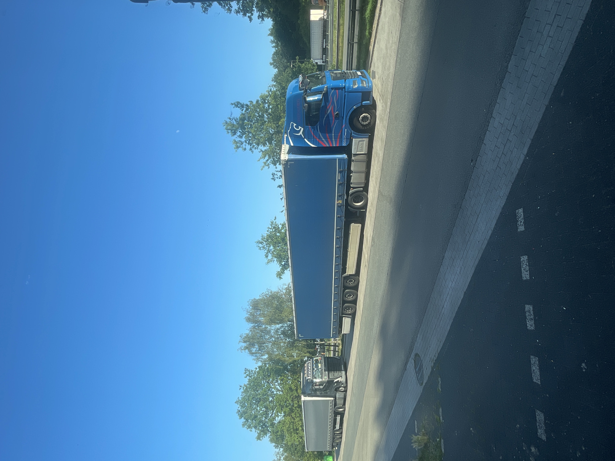 The image shows a roadside view in Ganderkesee, Germany. Two semi-trailer trucks are parked or stopped on a paved area next to a road. The truck on the left is dark gray and has "TRANSMAREK" written on its side. The truck on the right is bright blue with a graphic design on the cab and a blue trailer. The sky is clear and blue with a faint crescent moon visible. Trees and greenery line the background. The foreground shows a paved area with markings.
