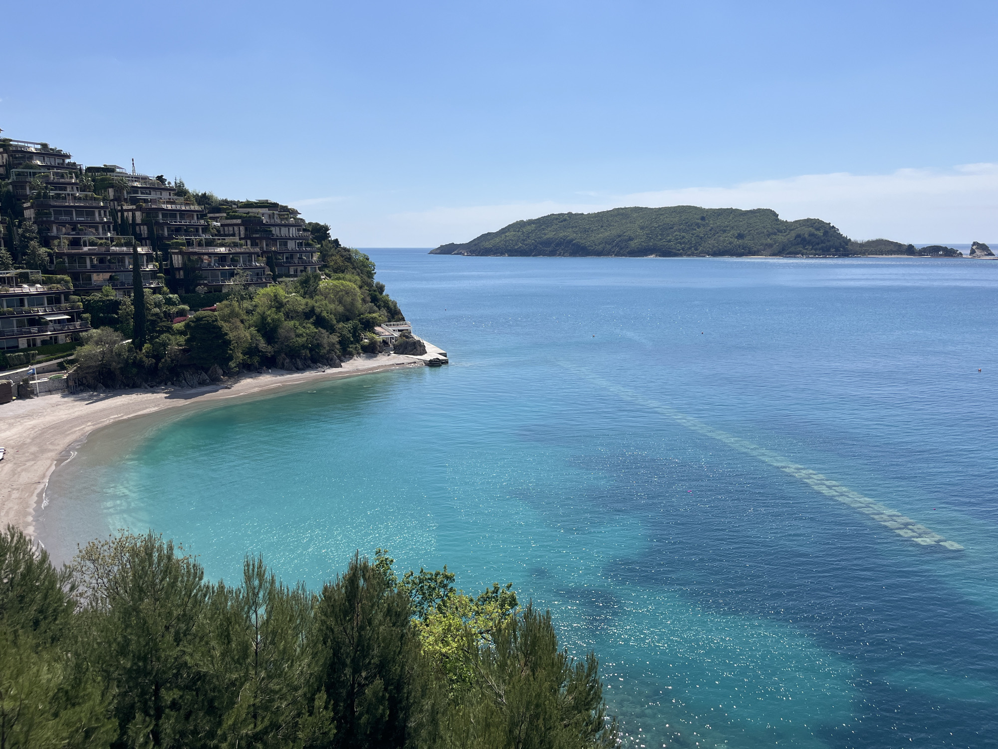 The image depicts a coastal scene with buildings nestled into a hillside on the left side of the frame. A small, sandy beach curves along the base of the hillside. The water in the bay is a turquoise color, transitioning to a deeper blue further out. An island covered in green vegetation is visible in the center of the frame. An underwater structure stretches out into the sea, visible as a lighter, rectangular pattern. The sky is clear and blue. The location is the Aman Sveti Stefan resort in Montenegro. In the foreground are trees.