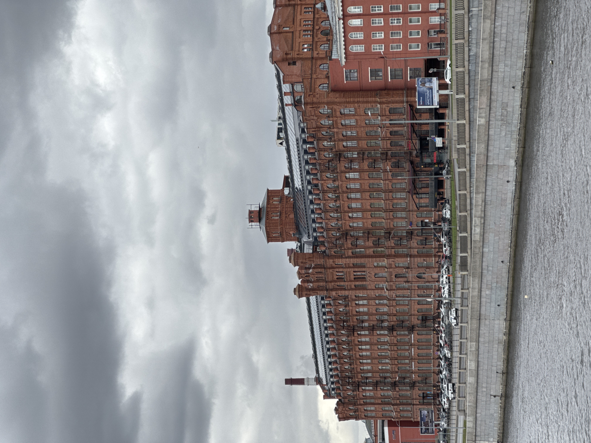 The image captures a panoramic view of a long, historic red brick building complex situated along a wide waterway in what appears to be Saint Petersburg, Russia, under an overcast sky.
The main subject is a series of interconnected red brick buildings, characterized by their uniform architectural style with numerous windows of varying shapes (rectangular, arched) and decorative brickwork. Some sections of the buildings feature modern glass structures on their roofs, suggesting renovation or contemporary additions. Towards the left, a tall, reddish-brown industrial chimney rises from the complex. In the foreground, a paved embankment with a metal railing separates the buildings from the tranquil, grey water of the river or canal. Several white cars are parked neatly along the embankment, indicating an urban setting. Small, dark shapes, possibly birds, can be seen on the water.
The weather is distinctly overcast, with a sky filled with various shades of grey clouds, suggesting a muted, possibly cool, daytime. The overall scene conveys a sense of calm and historical grandeur.
Visible text on a billboard includes Cyrillic words such as "Аристократ" (Aristocrat) and "Безопасно" (Safely/Securely), further confirming the Russian location. A small figure, likely a person, is standing near one of the billboards, providing a sense of scale. The activity depicted is that of everyday urban life by the water, with parked cars and the grand architecture dominating the view.