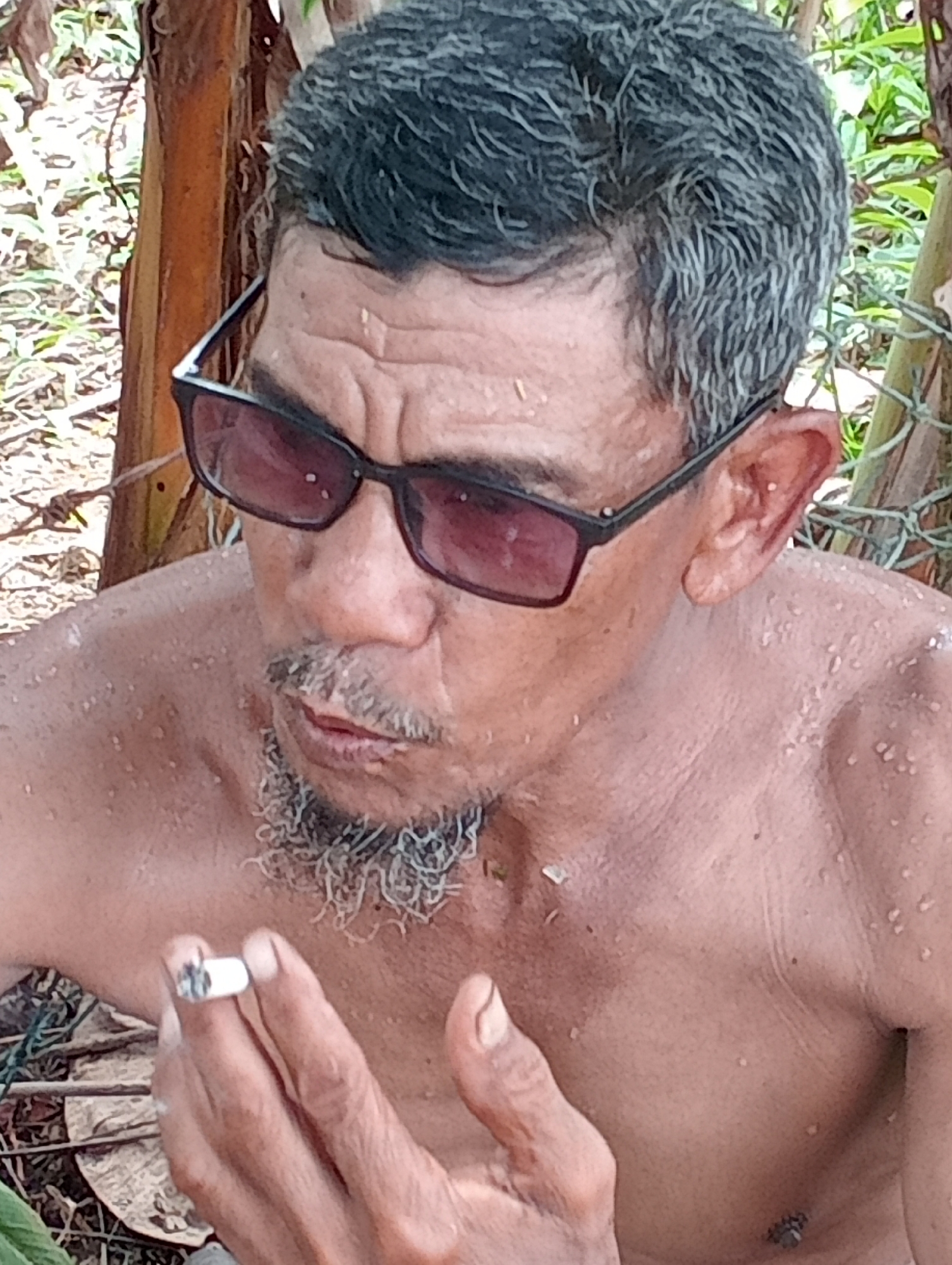 A man with gray hair, wearing dark sunglasses, is shown from the chest up, holding a cigarette in his right hand. He has a sparse gray beard and appears sweaty. He is standing outside in a natural environment with tree trunks and foliage visible in the background. The city and specific location are not discernible from the image.