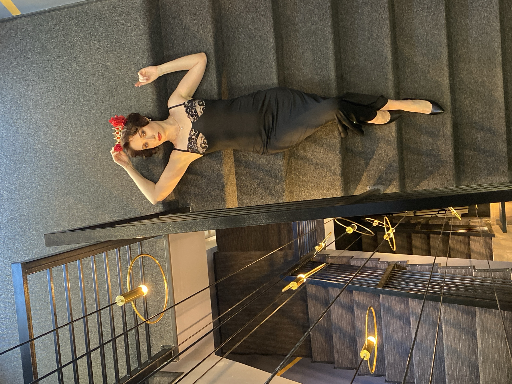 A woman in a black dress and red floral headpiece lies on a gray staircase. The staircase is part of a modern interior, featuring a metal railing with integrated lighting fixtures. The lights are ring-shaped, and the staircase appears to descend multiple floors. The location is in Yekaterinburg, Russia.