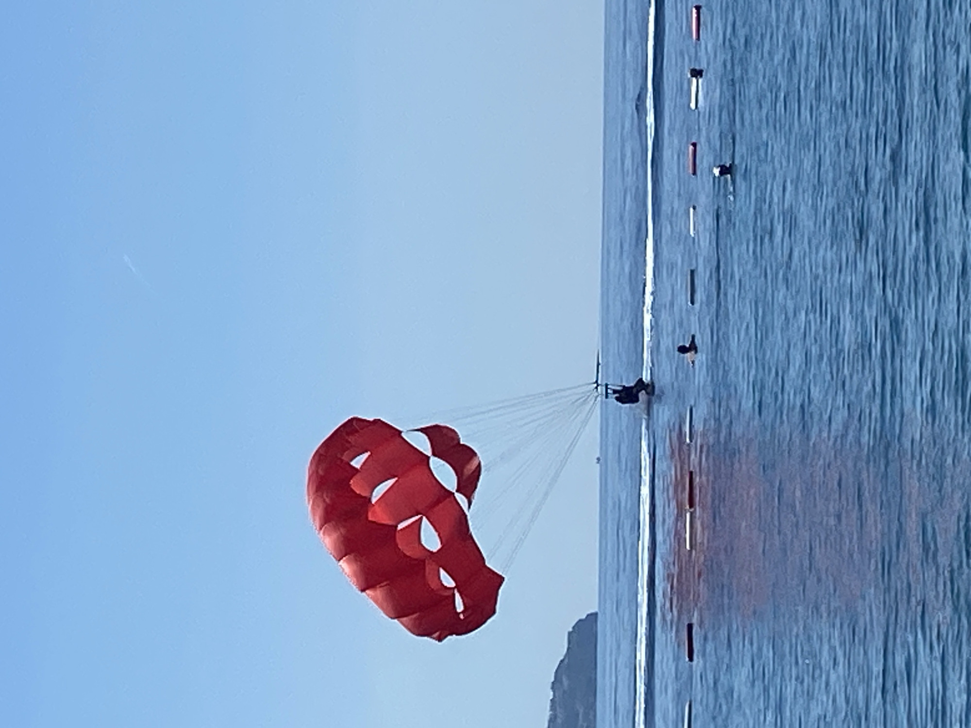 The scene depicts a bright, clear day in a coastal area of Budva, Montenegro, focused on water activities. A large, red parasail with multiple white, tear-drop-shaped cut-outs is centrally positioned, descending towards the water. A single individual, dressed in dark clothing, is suspended in a harness beneath the parasail, just above the water's surface, creating a wake suggesting movement by an unseen towing vessel. The calm blue sea features a delineated swimming area marked by a line of floating buoys, alternating between white, grey, and red. Within this area, a prominent red reflection from the parasail is visible on the water. Three individuals are observed swimming in the water near these buoys; one is to the far left of the buoys, another wears a white cap in the center, and a third is partially visible to the right. In the background, a distant landmass, possibly a vegetated hill or cliff, is visible on the left horizon, with the open sea extending to the right. A faint, white contrail is discernible in the upper right quadrant of the clear blue sky.