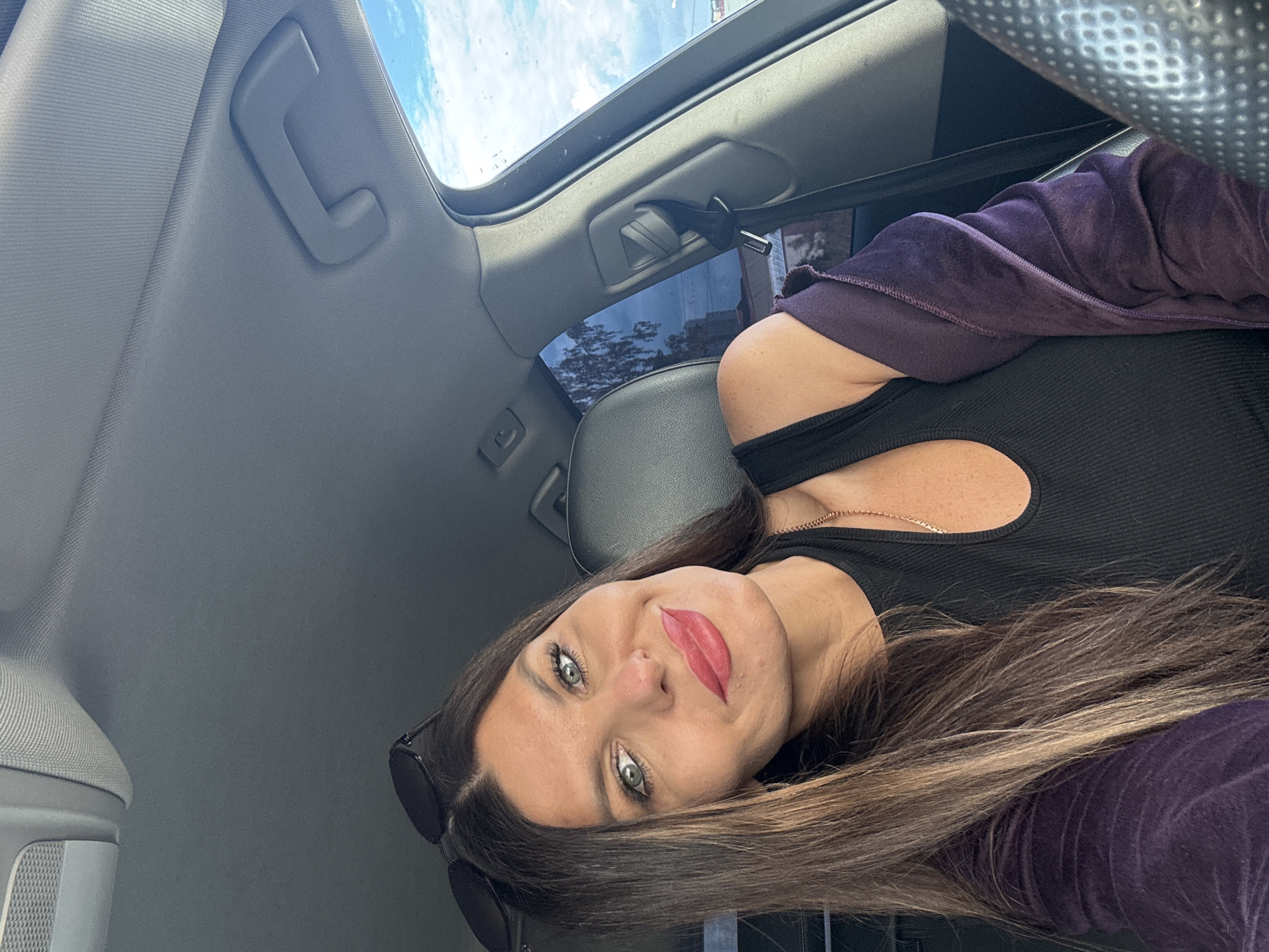 The media file is a selfie taken inside a car. The primary subject is a woman with long brown hair and striking green eyes, her face angled towards the camera. She is wearing a black, fitted top with a cutout detail and a purple velour jacket draped over her shoulders. Her lips are painted a vibrant red. She is also wearing sunglasses on top of her head and a delicate necklace.
The setting appears to be daytime, indicated by the bright sky and clouds visible through the car's sunroof. The interior of the car is grey and appears to be a typical passenger vehicle. The woman is likely the driver or a passenger. The overall mood conveyed by her expression and makeup is confident and perhaps playful.
There is no visible text in the media file. The provided location context of Chaikovsky, Russia, cannot be confirmed or denied by visual cues within the image itself.