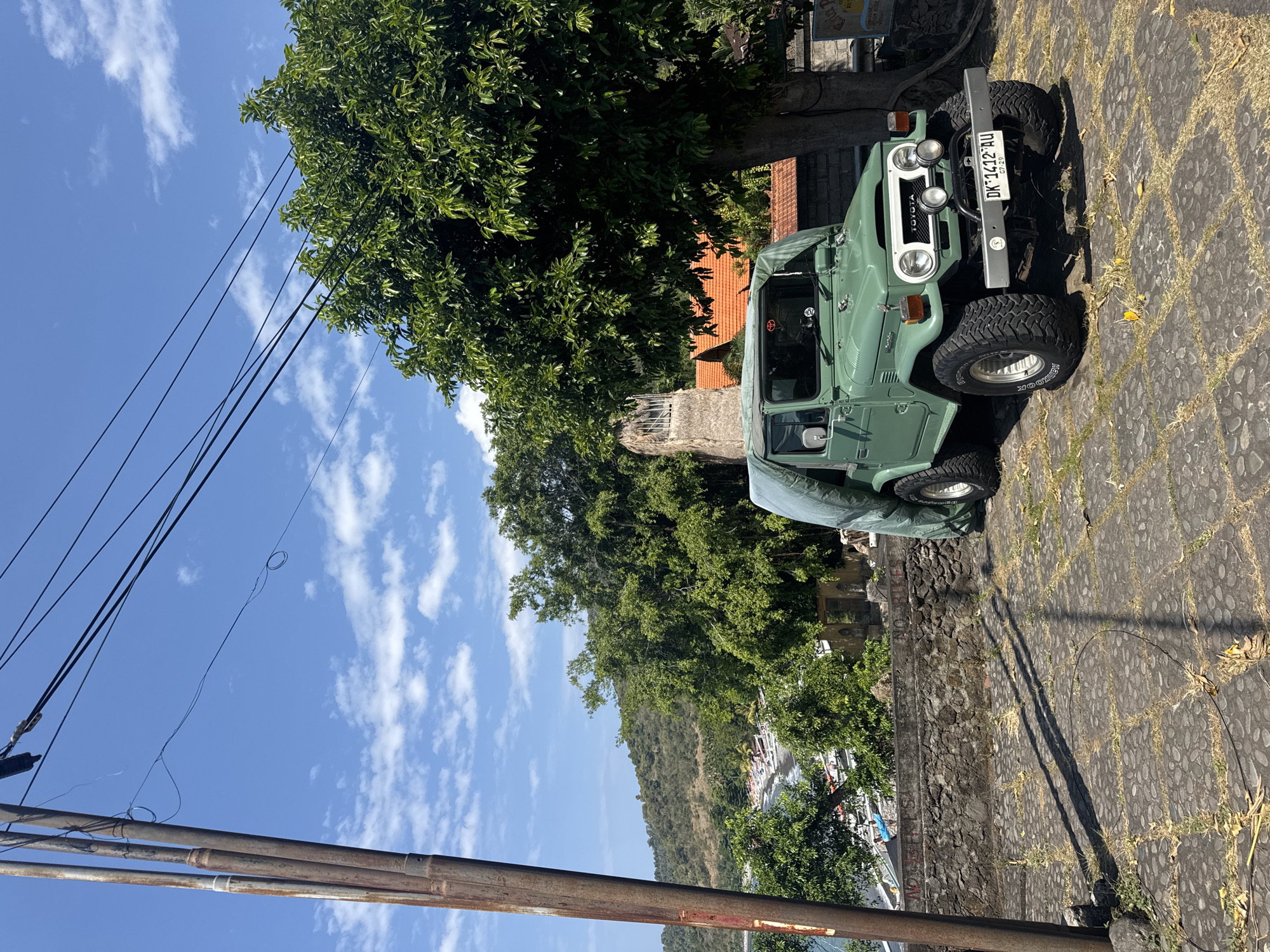 In Abang, Indonesia, a vintage green Toyota Land Cruiser is parked on a stone-paved area. The vehicle is partially covered by a light green tarp. It is situated outdoors, with a lush green tree to its right and a stone wall behind it. In the background, there's a glimpse of a body of water with boats, hills covered in vegetation, and a clear blue sky with scattered clouds. Power lines are visible stretching across the sky from a utility pole on the left side of the frame. The license plate of the vehicle reads "DK 1412 AU".
