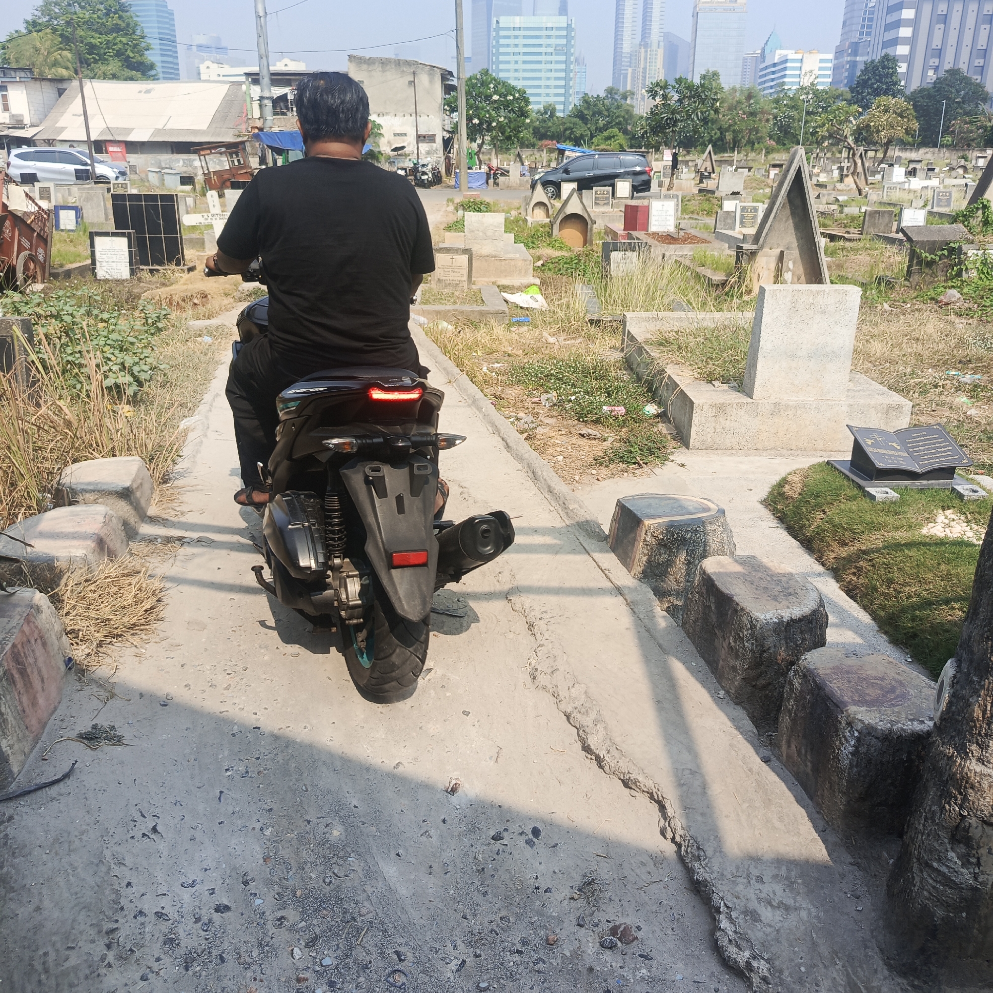 A man is riding a scooter on a paved path through a cemetery. The cemetery is filled with gravestones of various sizes and styles. In the background, there are buildings, including modern skyscrapers, and trees. The sky is clear and the day appears to be sunny. The man is wearing a black t-shirt and black pants. The scooter is black and has a red taillight. The path is concrete and has some cracks and uneven sections. There are also concrete blocks and some vegetation along the sides of the path. The overall scene depicts a contrast between the urban environment and the solemnity of the cemetery.