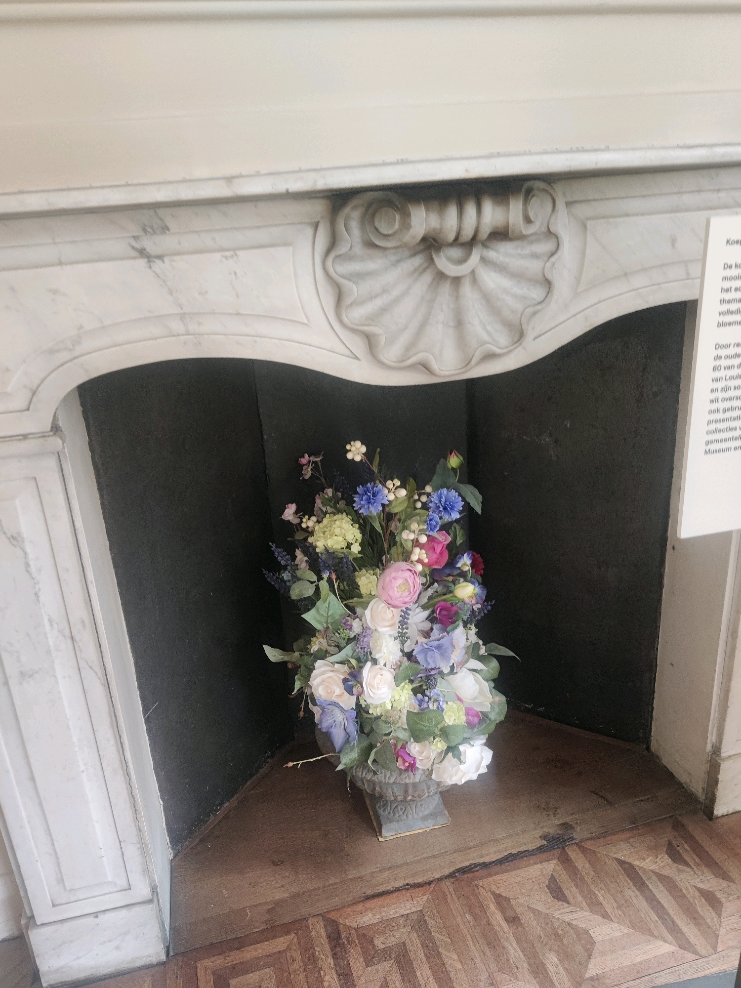 The image shows a floral arrangement in a vase placed in front of a fireplace. The fireplace has a marble mantelpiece with ornate carvings. To the right of the fireplace, a white sign with text in Dutch is visible. The floor is made of wooden parquet. The floral arrangement consists of various flowers in shades of white, pink, purple, blue, and green, with some greenery. The vase is a decorative stone-colored urn. The overall scene appears to be indoors, possibly in a museum or historic building.