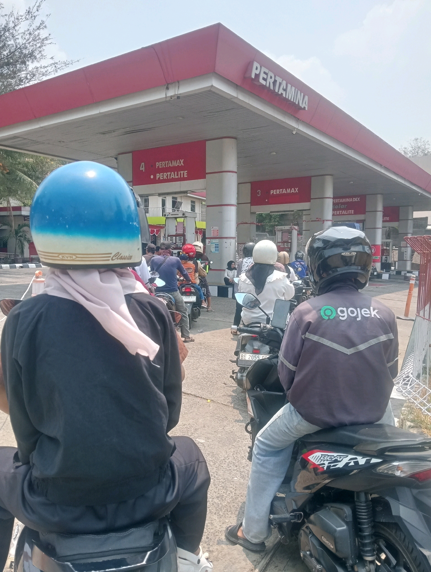 The image depicts a queue of motorcyclists at a Pertamina gas station in Bandar Lampung, Indonesia. The station features a prominent red roof with white columns and signage indicating different fuel types like "Pertamax" and "Pertalite." In the foreground, two individuals on motorcycles are waiting in line. The person on the left wears a blue and white helmet and a black jacket with a light-colored scarf. The person on the right wears a black helmet and a dark grey jacket with the "Gojek" logo on the back. Several other motorcyclists, some with helmets and some without, are visible further down the queue. The environment appears to be a typical urban setting with a paved road and a clear sky. The presence of multiple motorcycles suggests it is a busy time at the gas station.