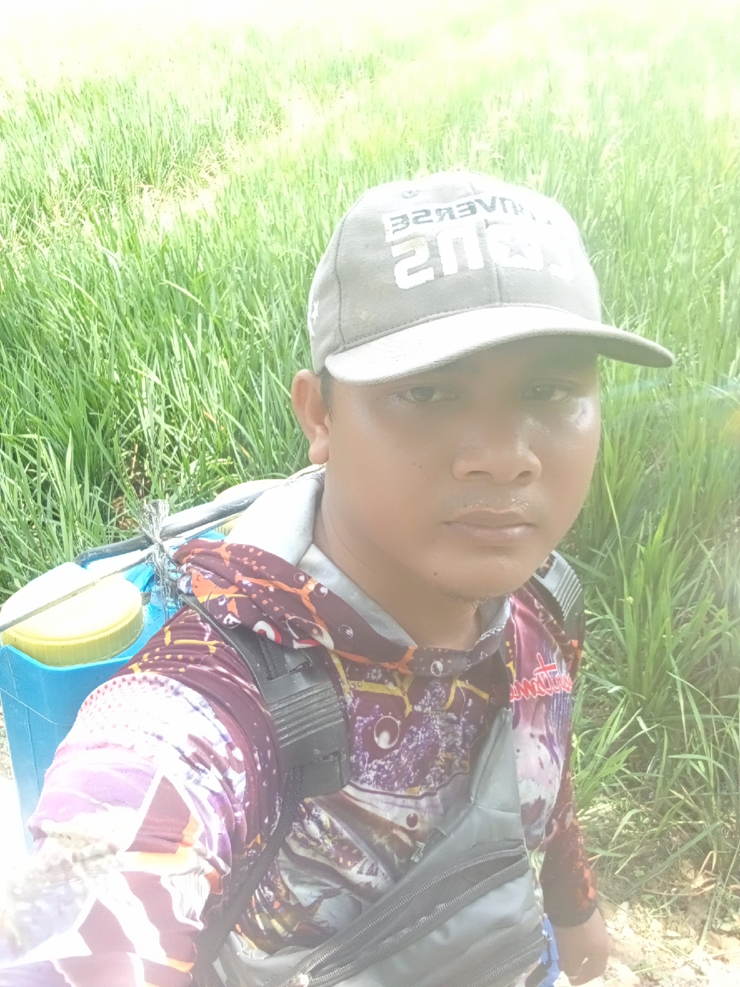The image is a selfie of a young man wearing a gray baseball cap with white text on it, a long-sleeved shirt with a colorful abstract pattern, and a black fanny pack worn across his chest. He is carrying a blue backpack with a yellow lid on his back. The man is standing in a grassy field. The overall lighting is bright and washed out, suggesting it is daytime. The location is identified as Sri Mulyo, Indonesia.