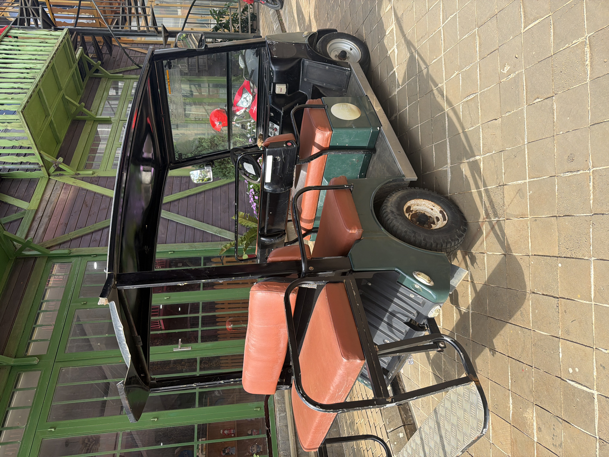 A multi-passenger utility vehicle, resembling a golf cart, is parked on a tiled outdoor surface in Măng Đen, Vietnam. The vehicle is predominantly dark green and black, featuring a hard roof, a windshield, and side mirrors. Its interior is fitted with tan or brown padded seats, including a front row and additional seating in the rear. A metal diamond plate step is visible at the back of the vehicle. Through the vehicle's front window, a red scooter or motorbike can be seen parked nearby. Small purple flowers add a touch of color near the driver's side.
The background features a building with a distinctive architectural style, characterized by prominent green wooden frames and large glass panels, suggesting it might be a cafe, restaurant, or resort structure. Dark wooden siding is also visible on parts of the building. The ground is paved with light-colored, square tiles, showing visible grout lines and some wear. Long, distinct shadows are cast across the tiled ground, indicating bright sunshine and suggesting either morning or late afternoon. No people are present, and no discernable text is visible on any objects.