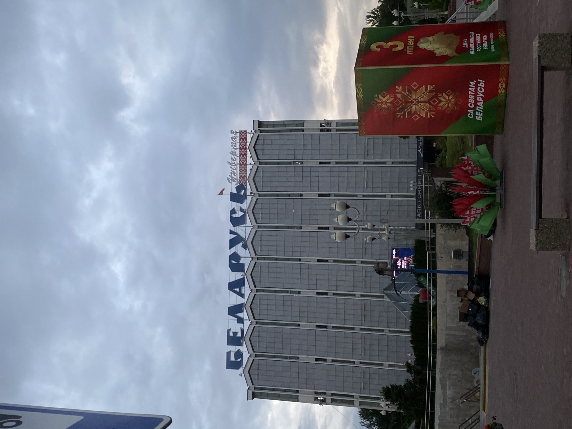 This media file captures a view of the "Belarus" department store in Minsk, Belarus, under a cloudy sky. The building's facade is predominantly glass and concrete, with large arched sections across its upper levels. A prominent blue sign with the word "БЕЛАРУСЬ" (Belarus) in large block letters is visible at the top of the building. To the right of the building, there's a large, decorative stand with red and green panels, featuring a large snowflake motif and text in Belarusian. One panel reads "3 ліпеня" (July 3rd), another displays a map of Belarus, and the main message proclaims "СА СВЯТАМ, БЕЛАРУСЬ!" (Happy holiday, Belarus!). This suggests the image was taken around Belarus' Independence Day, celebrated on July 3rd.
In the foreground, a pile of garbage bags and cardboard boxes is present on a paved area, next to some decorative red and green elements that appear to be artificial flowers or ribbons. The time of day is likely late afternoon or early evening, given the soft, diffused light from the overcast sky. The overall atmosphere is somewhat subdued due to the weather and the presence of the trash, contrasting with the celebratory decorations. Streetlights are visible on poles, indicating an urban setting. The image is oriented vertically.