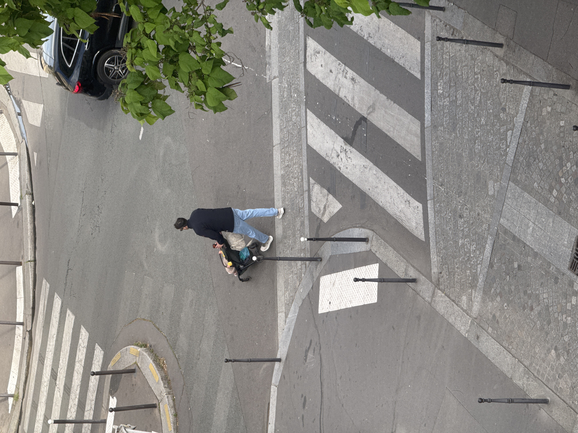 The image depicts an overhead view of a street scene in Paris, France. A person wearing a dark blue jacket and light blue jeans is pushing a stroller. They are walking on the street near a pedestrian crosswalk with black and white stripes. A black car is parked at the edge of the street near some green foliage. There are black bollards along the street and sidewalk edges. The ground is paved with a mixture of asphalt and stone.