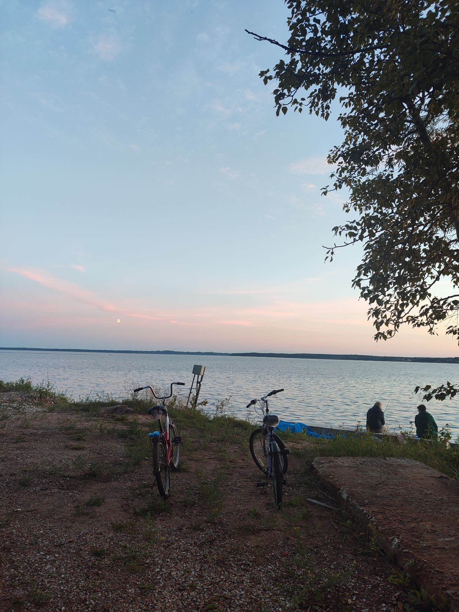 The image depicts a tranquil lakeside scene at dusk. Two bicycles, one red and one blue, are parked on a gravelly shore. In the background, a calm body of water stretches to the horizon, with a faint pinkish hue in the sky indicating the setting sun. A crescent moon is visible in the sky. Several people are seated by the water's edge, appearing to be fishing. The environment is natural, with some vegetation along the shoreline and a large tree with dark leaves partially obscuring the upper right corner of the frame. The overall mood is peaceful and serene.