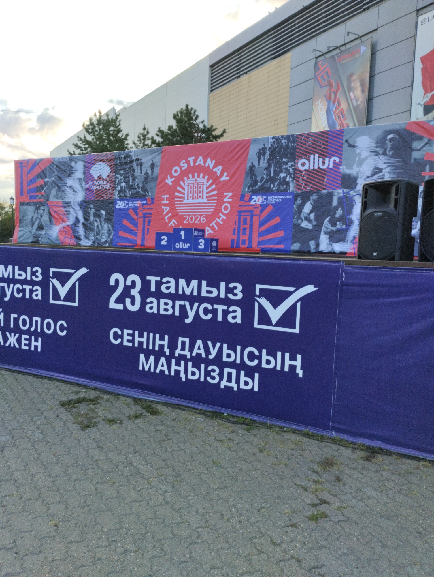 The image shows a large blue banner with white text in both Kazakh and Russian, displayed outdoors, likely in front of a building with a large facade. The banner features a checklist icon with a checkmark. The text on the banner appears to be related to an event, possibly a marathon or a voting process. Specifically, it includes the date "23 августа" (August 23rd) in Russian and "23 тамыз" in Kazakh, suggesting an event taking place on that date. The phrase "Сенің дауысың маңызды" (Your voice is important) is also visible in Kazakh, indicating a civic or participatory element.
Above the blue banner, a red banner with various images and text is visible. This banner seems to be advertising a "Kostanay Half Marathon" with "2026" mentioned, and "World Athletics" branding. Images of runners and athletic imagery are present.
The overall setting suggests an urban environment, possibly a public space in the city of Kostanay, Kazakhstan, where a promotional event or a public announcement is taking place. The ground appears to be paved with interlocking stones. Two large black speakers are visible on the right side of the image, suggesting an audio component to the event. The sky in the background shows some clouds and trees can be seen to the left, indicating an outdoor setting.