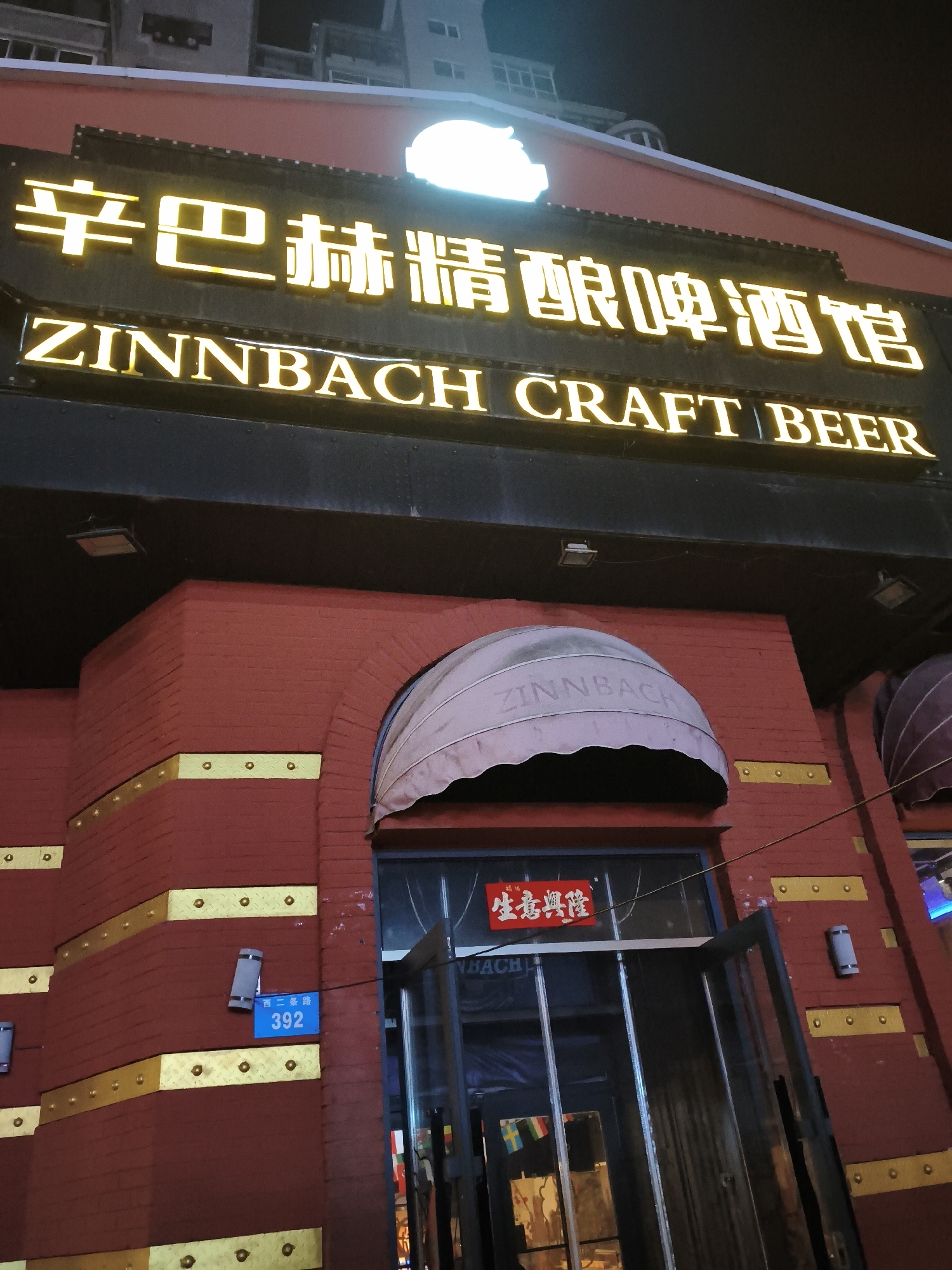 The image shows the exterior of a Zinnbach Craft Beer establishment at night. The building features a red brick facade with gold decorative elements. Above the entrance, a large illuminated sign displays "ZINNBACH CRAFT BEER" in English, with Chinese characters above it. There is an arched awning above the entrance, also bearing the name "ZINNBACH". A blue street number sign displaying "392" is visible on the brick wall. Inside the entrance, a red sign with Chinese characters reads "生意興隆", meaning "May your business flourish". The scene suggests a commercial establishment in an urban setting in Xi'an, China.