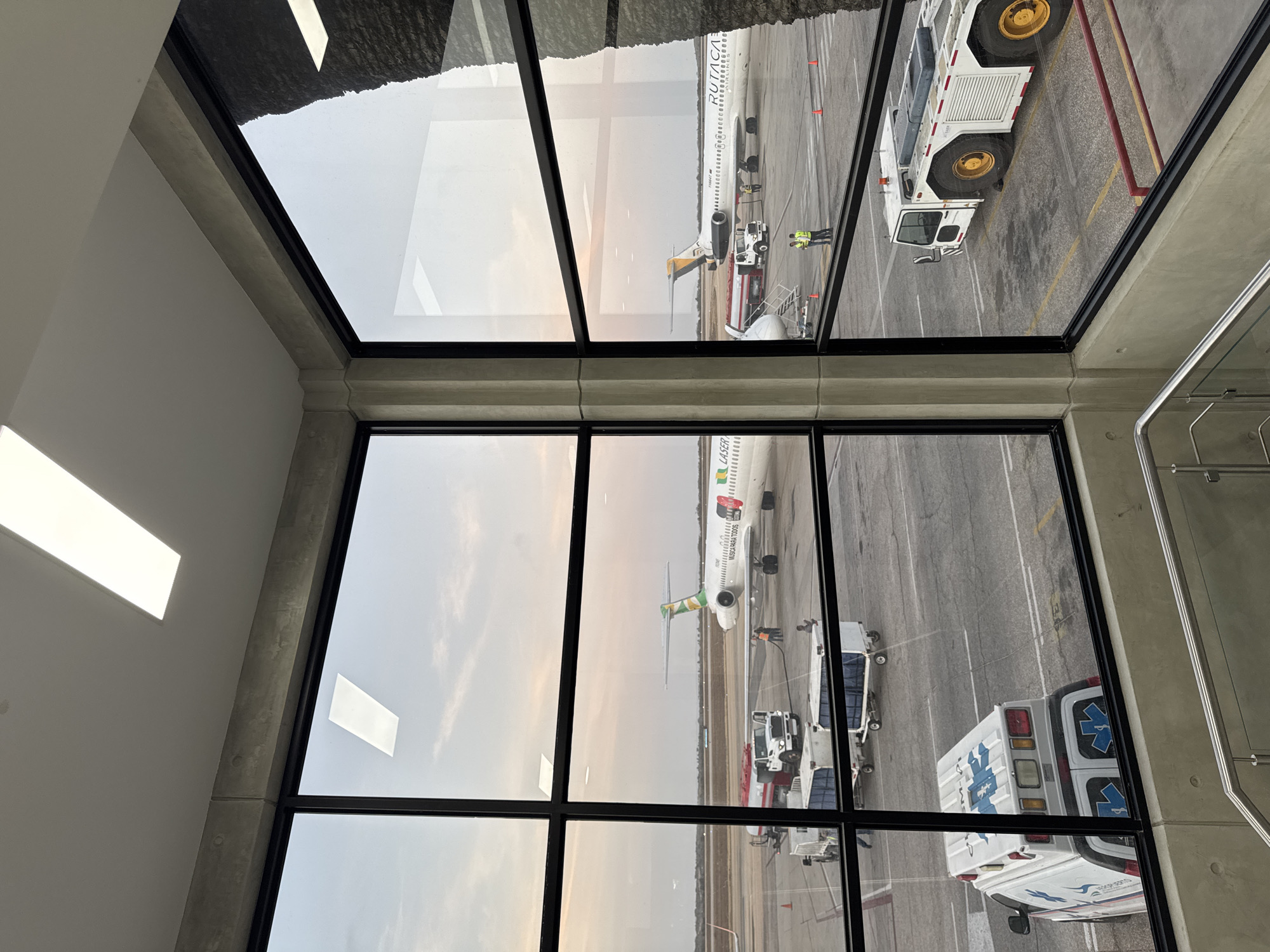 A view from inside a building, looking through a large window with black frames, reveals an airport scene. Two aircraft, one identified by the tail markings and "LASER" on the body and the other aircraft is identified with "RUTACA" painted on its side, are parked at the gate. Airport vehicles, including an ambulance and tug, are positioned near the planes. The sky appears overcast, with limited visibility. There are people servicing the aircraft. The location appears to be the airport in Cotoperíz, Venezuela, based on the presence of the airline names on the aircraft.