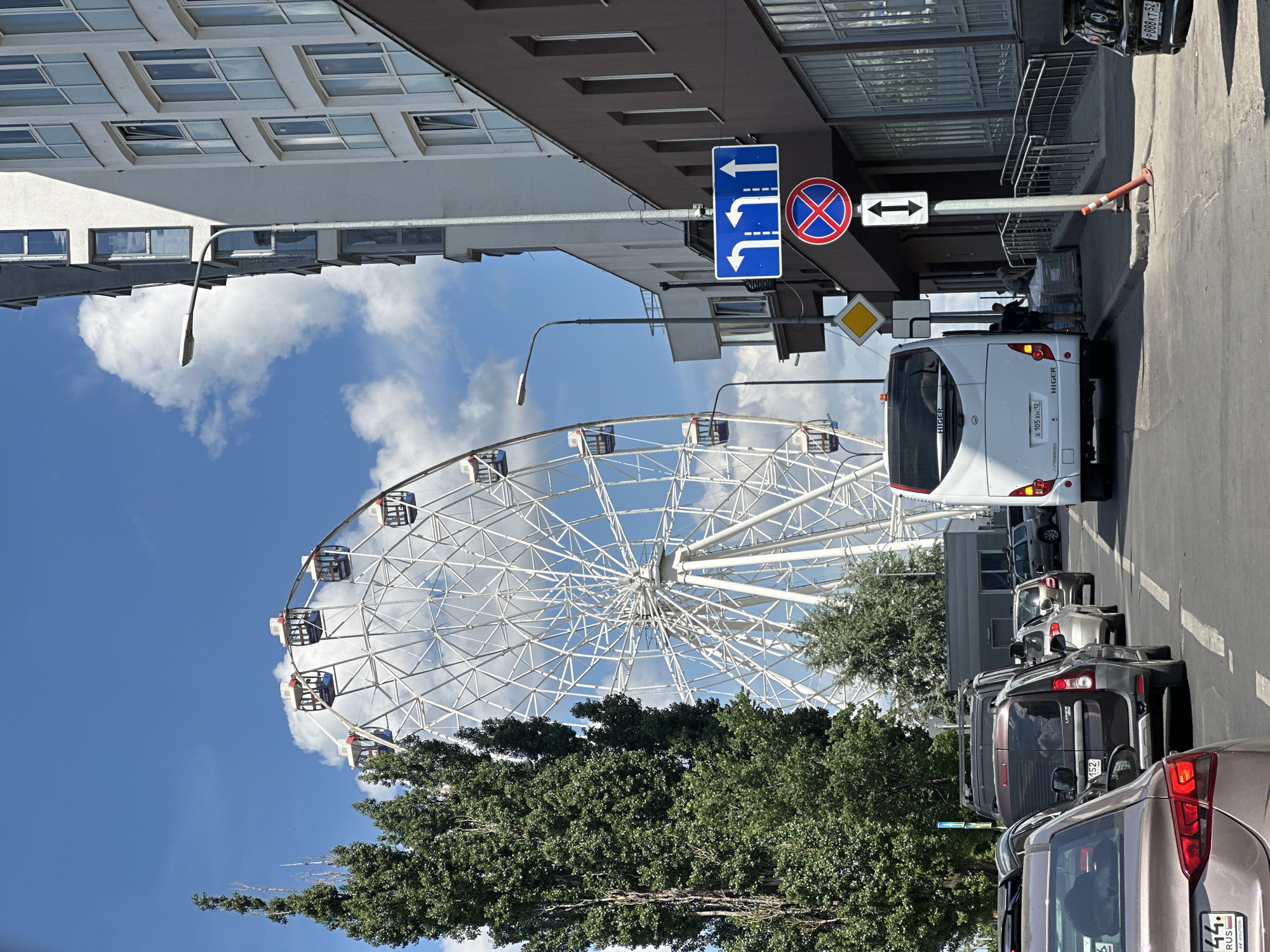 The image depicts a street scene in Nizhny Novgorod, Russia, dominated by a large white Ferris wheel in the background. A white bus with "HIGER" branding is stopped on the right side of the road, with two individuals near its rear, appearing to interact with a pallet of goods. Several cars are parked or moving in traffic on the road. A modern white building with numerous windows stands to the right of the Ferris wheel, casting a shadow. Traffic signs, including lane direction indicators and a "no stopping" symbol, are visible near the bus. Lush green trees are present on the left side of the frame, contrasting with the urban architecture. The sky is blue with scattered clouds.