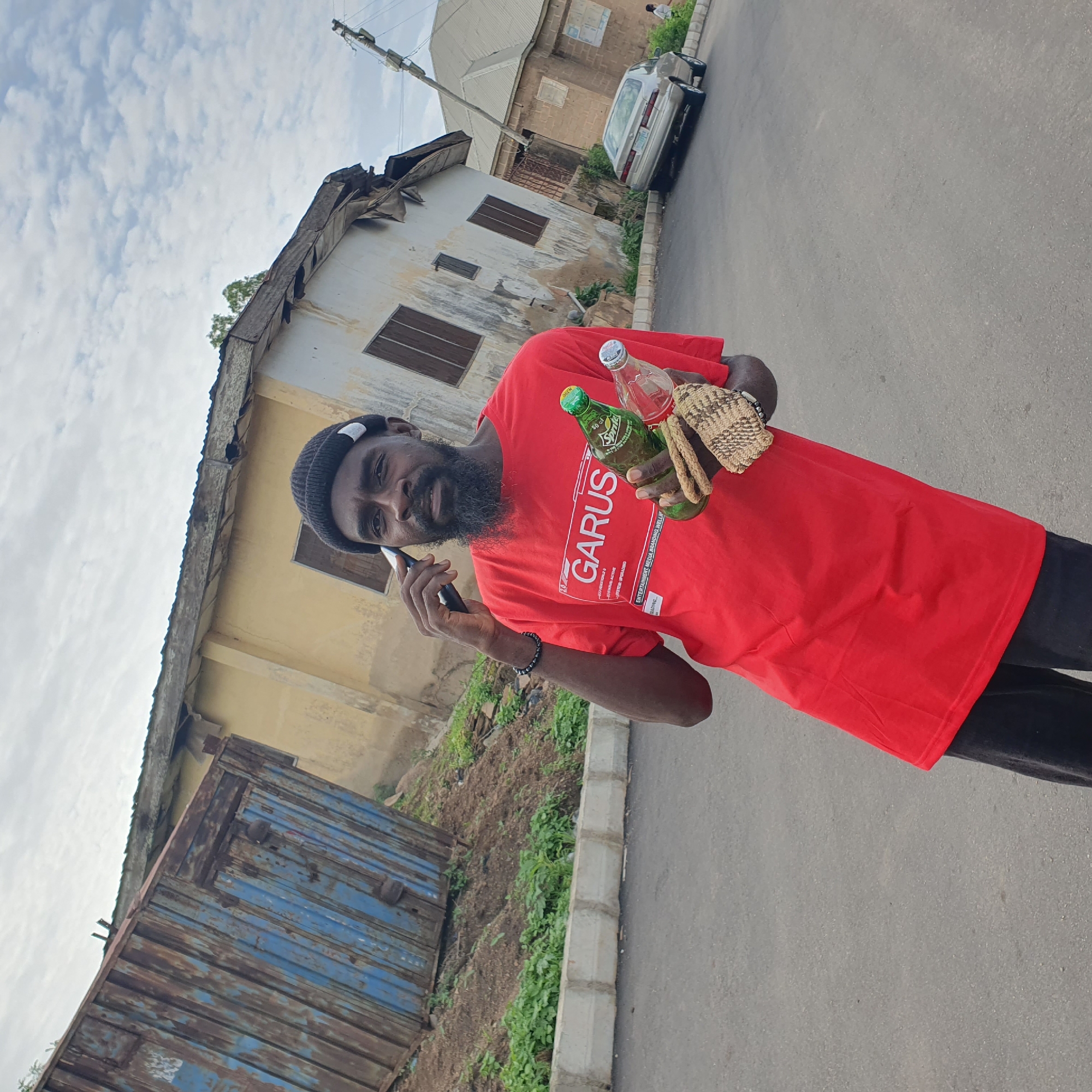 A man wearing a red t-shirt and a black beanie is standing on a paved road in Jos, Nigeria, holding two beverage bottles and a woven item. He is talking on a mobile phone. To his left, a large, rusty blue container is situated on a sloped, overgrown area. Behind him, a yellow, dilapidated building with boarded windows stands at an angle. Further in the background, a silver car is parked on the side of the road, and another building made of brick is visible. The sky is cloudy.