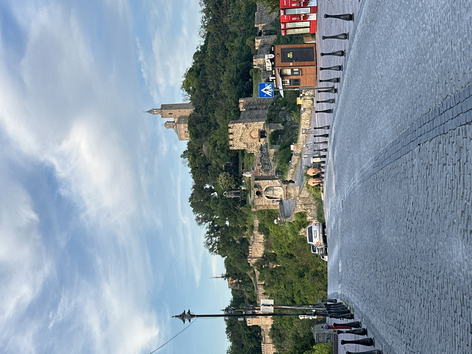 The image shows a view of Tsarevets fortress in Veliko Tarnovo, Bulgaria. The fortress is situated on a hilltop, surrounded by lush green trees. The foreground features a cobblestone street leading uphill towards the fortress. Several cars are parked on the street, and a few people are walking around. There is also a small ticket booth near the entrance to the fortress. The sky above is blue with some scattered clouds. The overall impression is a historic and scenic location.