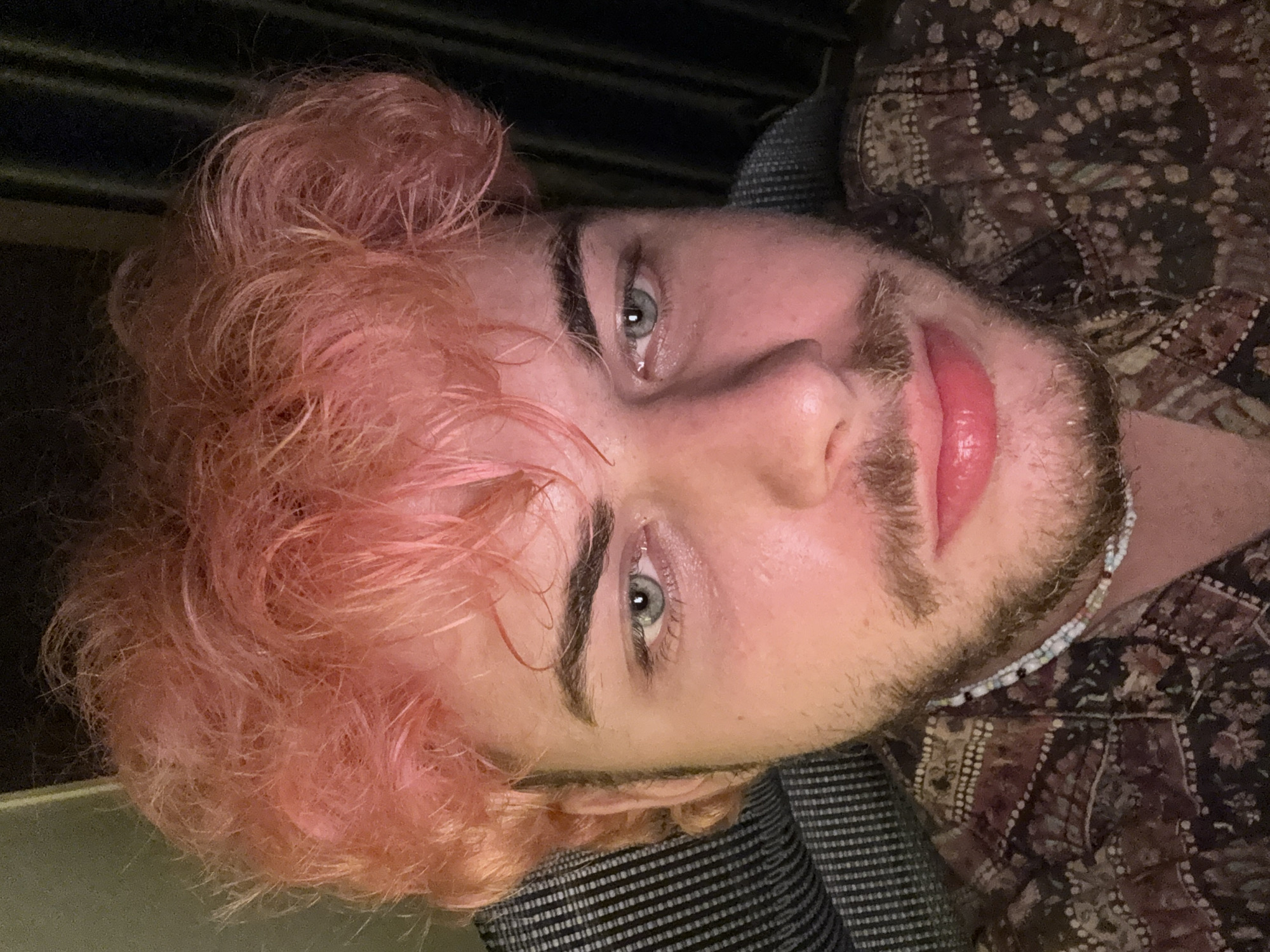 A man with pink hair and light eyes is looking directly at the camera. He has a slight beard. He is wearing a patterned shirt and appears to be sitting on a patterned seat. The background is dark and somewhat indistinct. There is nothing in the image to suggest a location in Đà Nẵng, Vietnam.