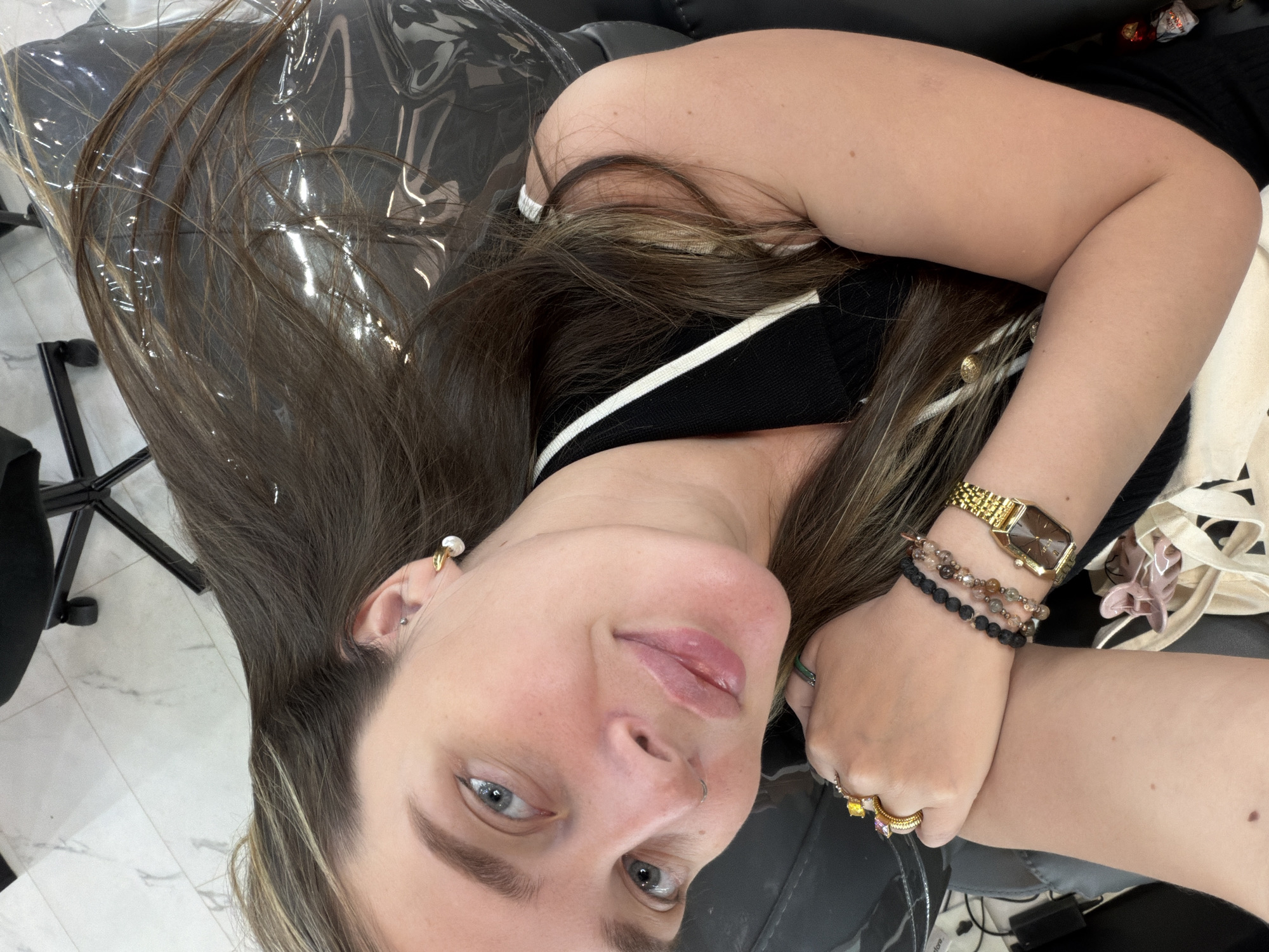 A woman lies on a black chair covered with clear plastic. She wears a black top with white straps, a gold watch, bracelets, and rings. Her long brown hair is spread across the chair. She has light skin, blue eyes, and a nose piercing. The setting appears to be a salon or a personal care space, with a glimpse of a marble floor. There is no indication of Moscow or Russia in the image.