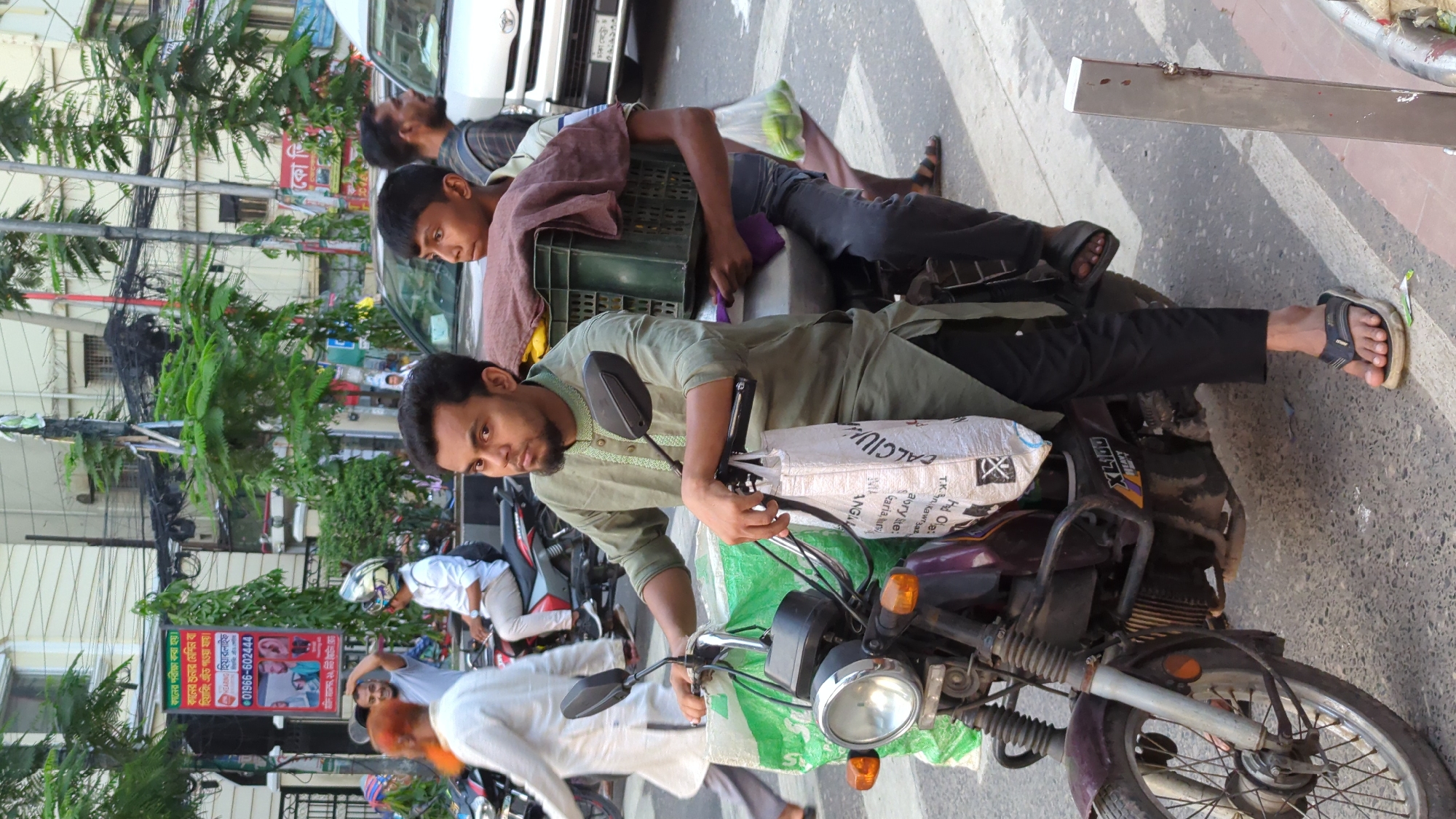 The image depicts a street scene in Dhaka, Bangladesh. A man in a green kurta is seated on a purple and silver motorcycle. A bag labeled "CALCIU" is placed on the motorcycle. A second man and a boy are sitting on what appears to be a parked car behind the motorcycle. A green crate is visible on the car's roof. Several people are walking on the sidewalk. A banner advertising "হিয়ারলাইফ" (HearingLife) with a phone number is visible. Trees line the street. The weather is sunny.